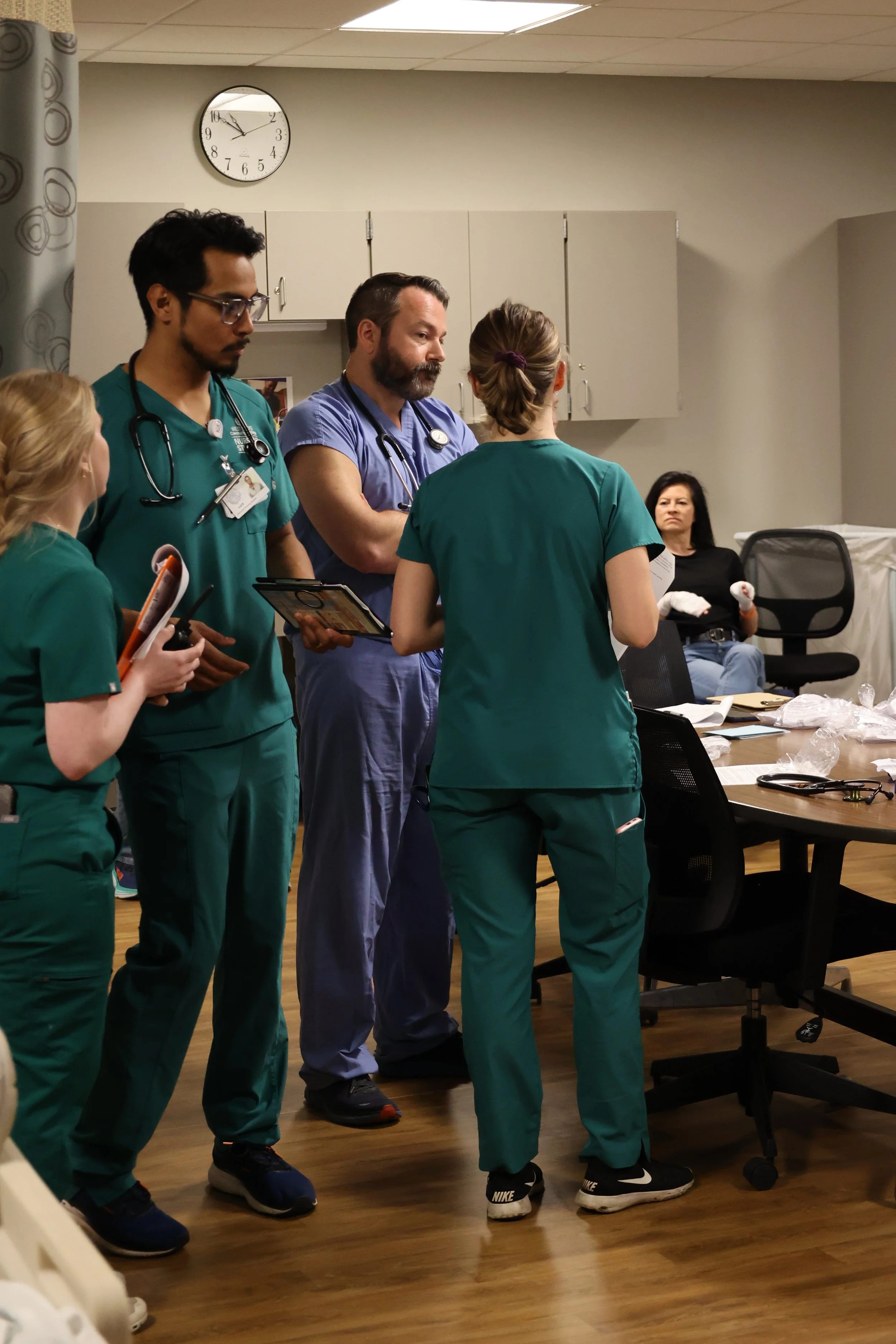 Healthcare professionals in discussion, wearing green and blue scrubs, in a hospital or clinic setting with medical equipment visible around a central table.