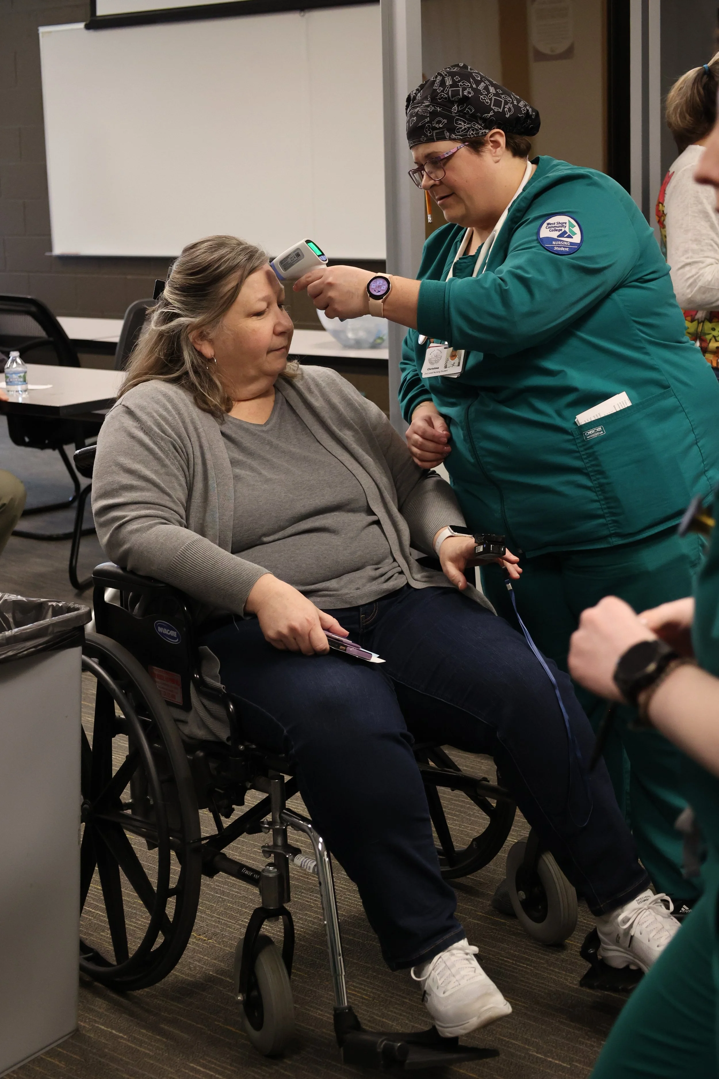A healthcare worker in green scrubs uses a digital thermometer to check the temperature of a woman sitting in a wheelchair.