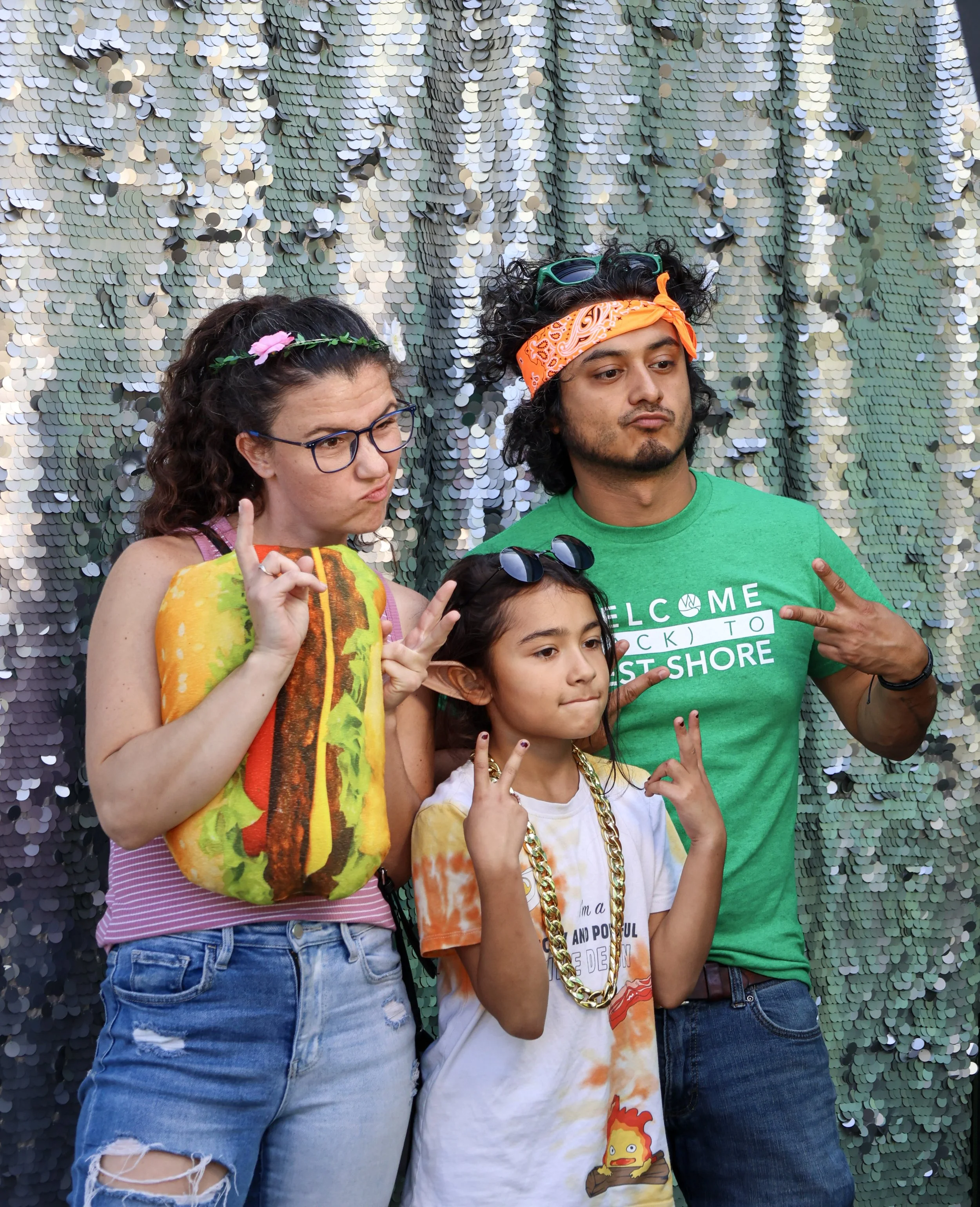 Three people posing in front of a shiny sequin backdrop, with the left person wearing a striped tank top and a hamburger-shaped pillow, the middle child wearing a T-shirt and sunglasses on the head, and the right person in a green T-shirt and orange bandana, all showing peace signs.