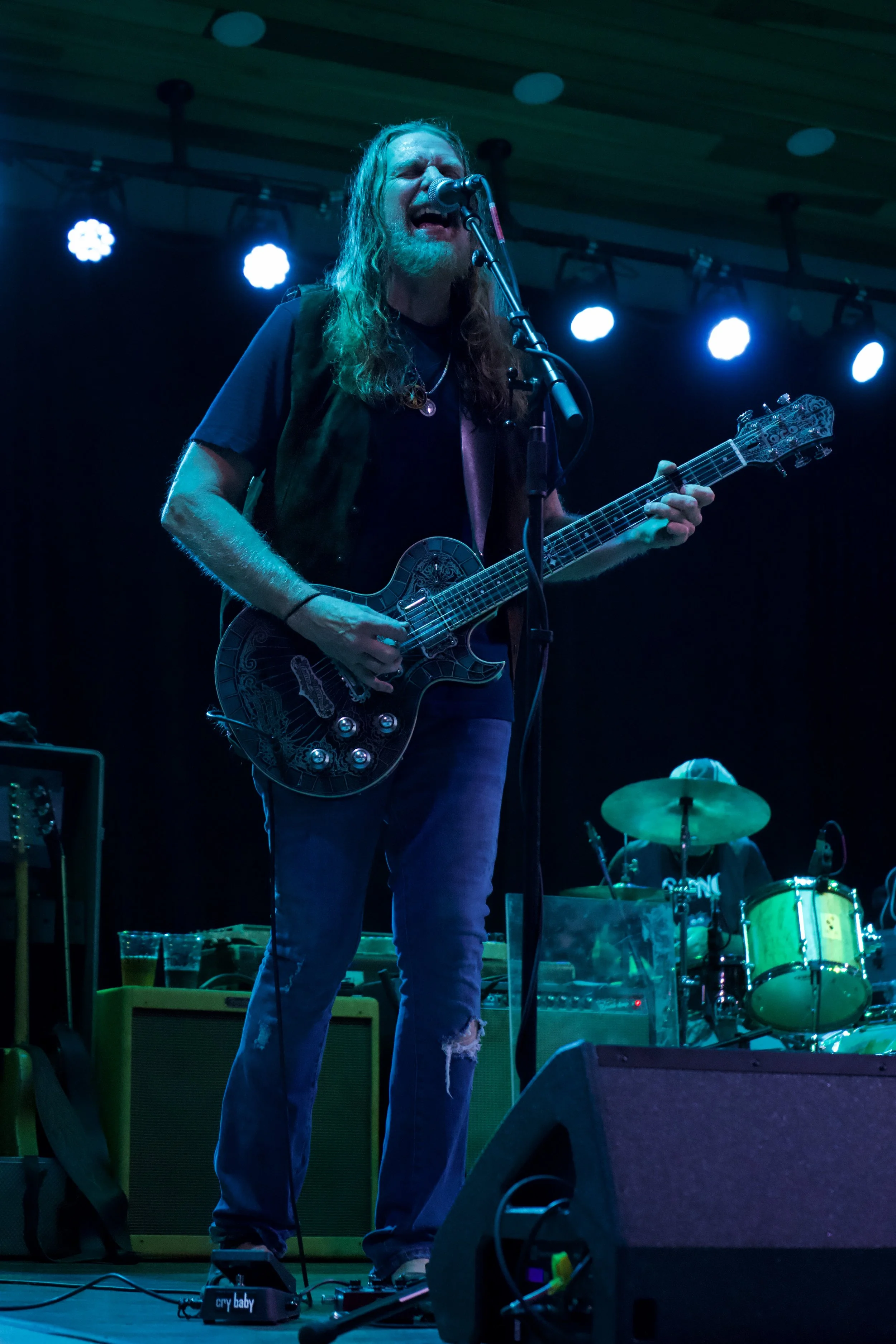Musician playing electric guitar and singing on stage with drummer in the background during a live performance.