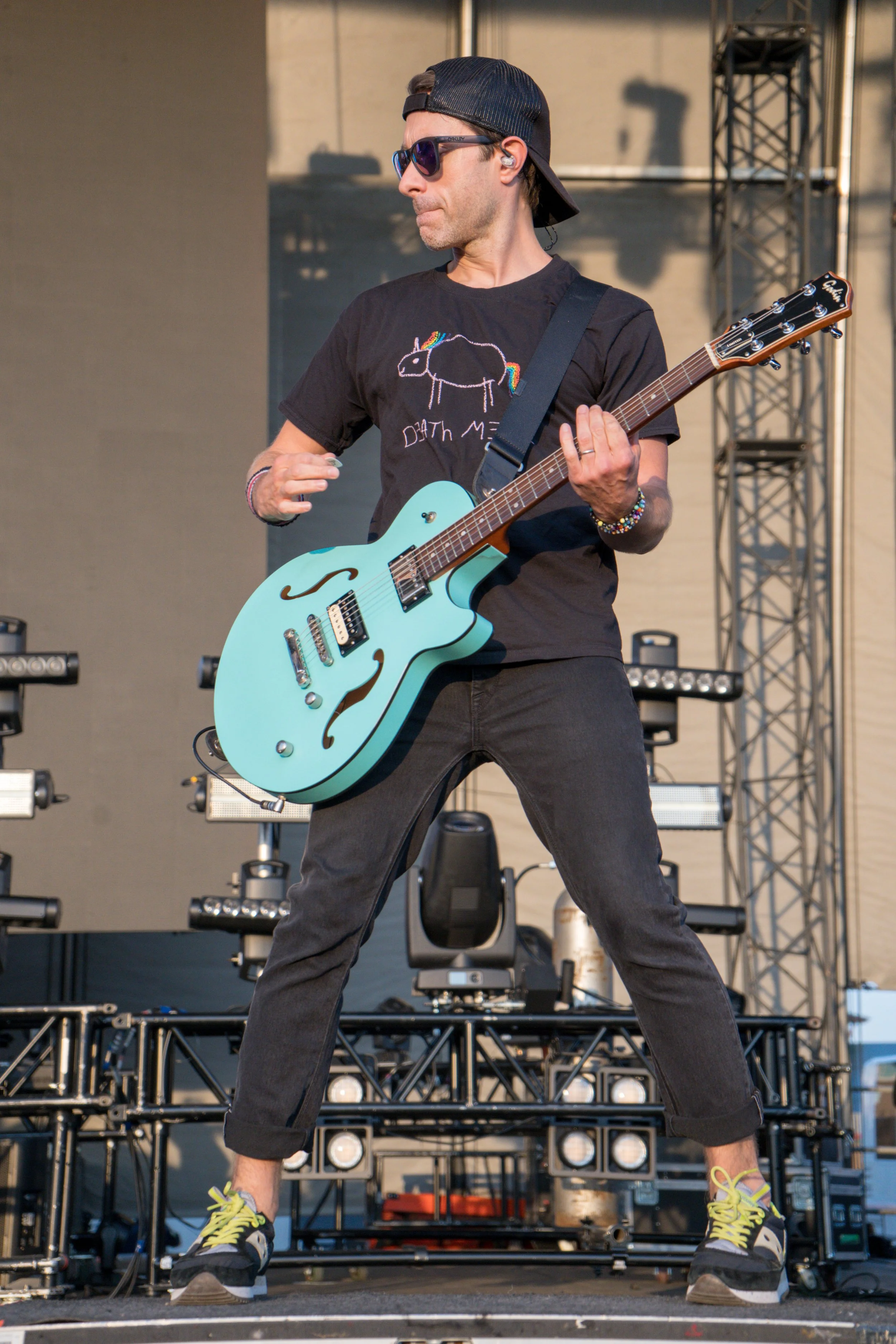 Musician playing light blue electric guitar on stage wearing black t-shirt, sunglasses, and backward cap.