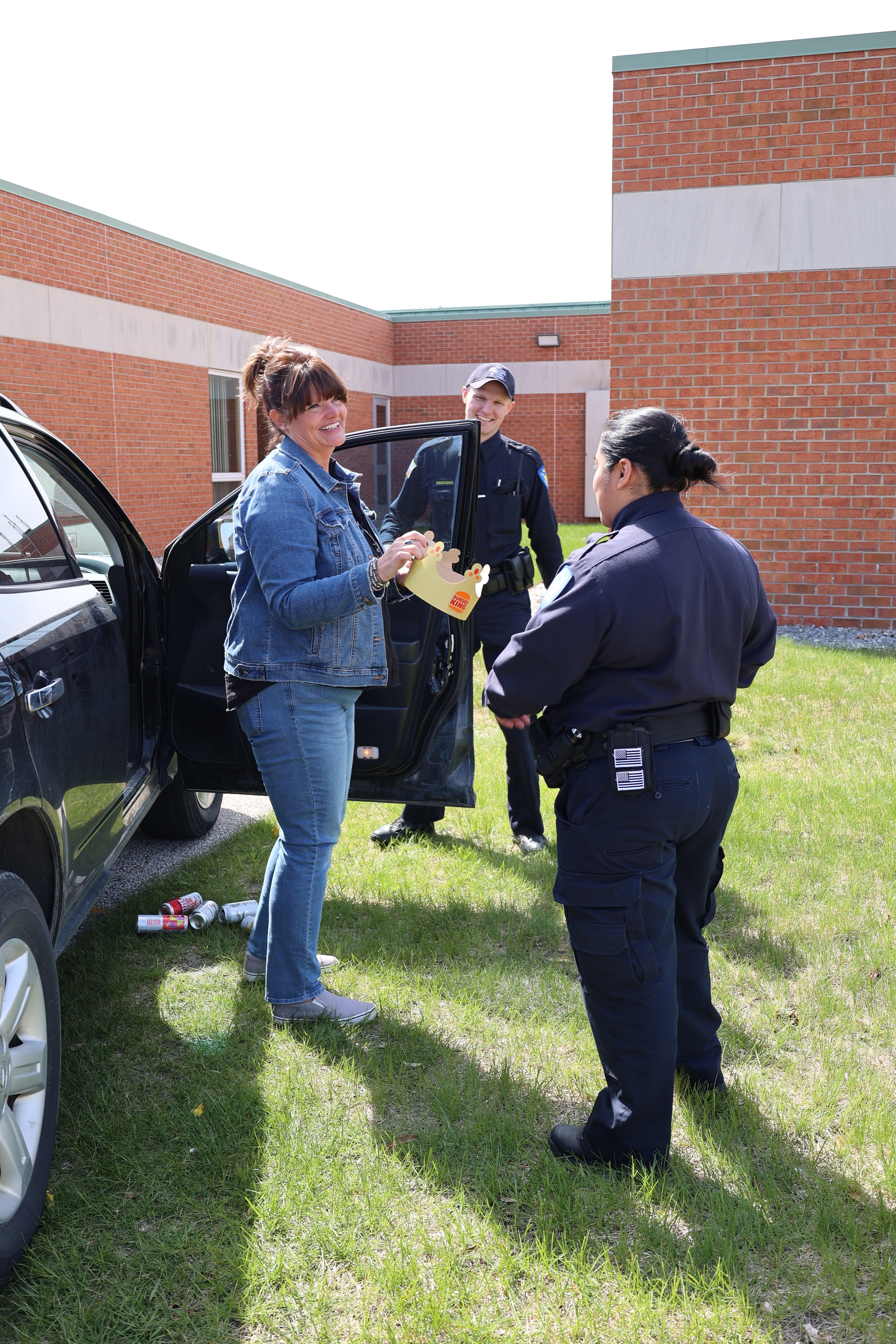 Two police officers talking to a woman holding a Burger King crown near a car with open door outside a brick building.