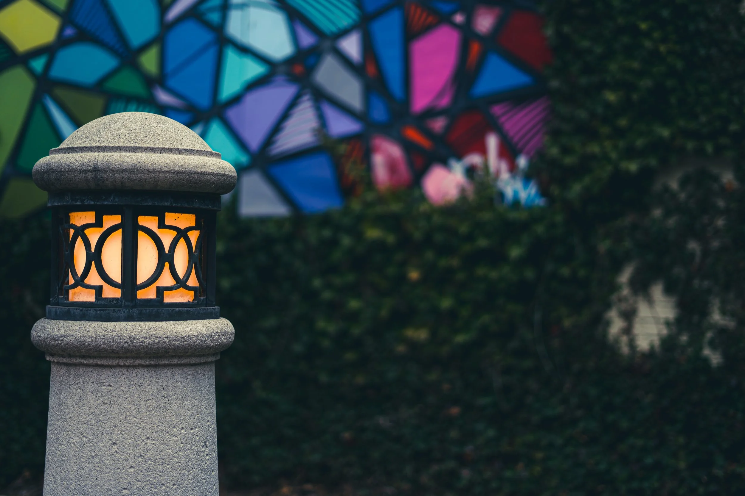 Lit outdoor lamp post in front of vibrant stained glass wall with greenery.