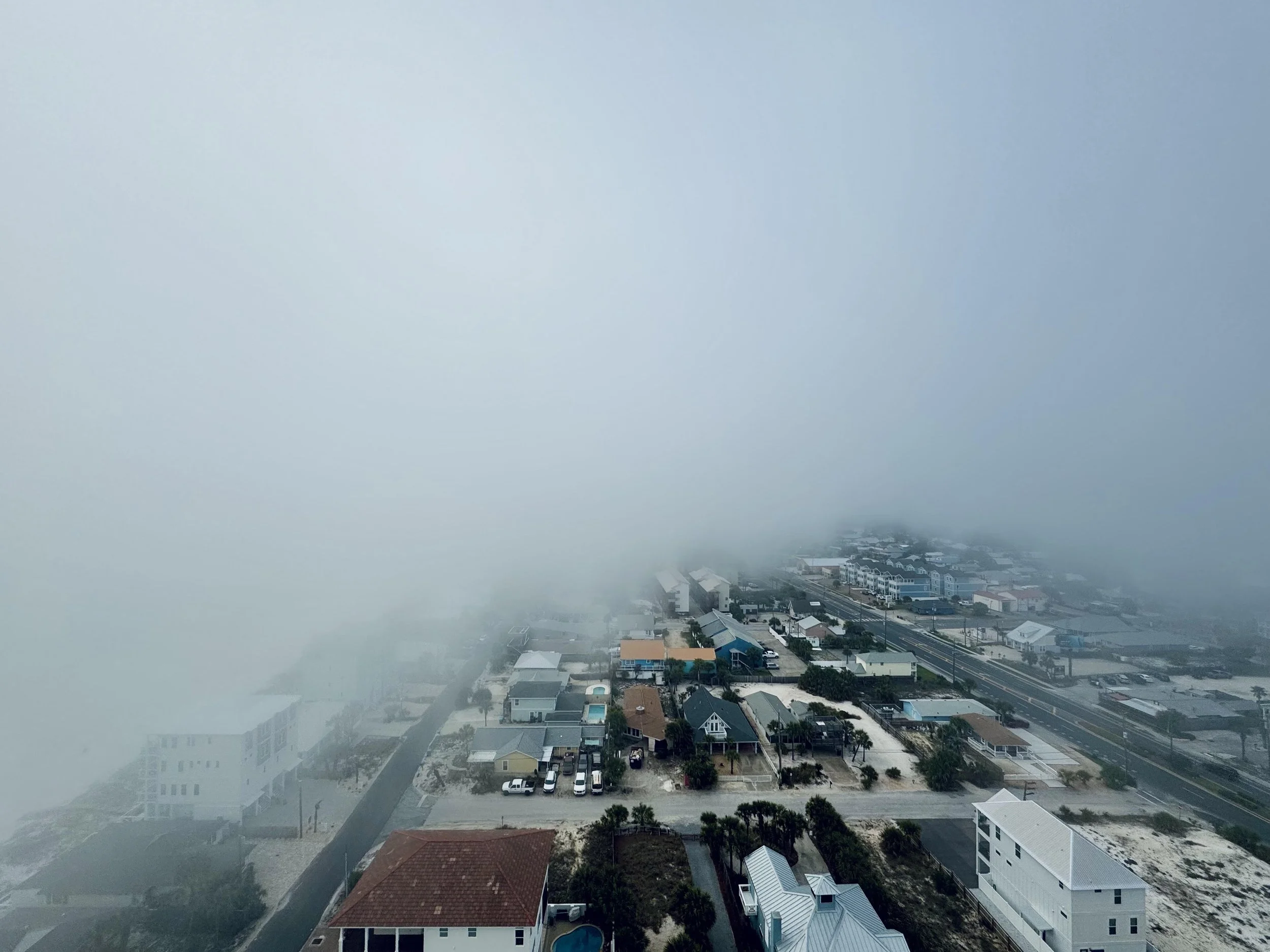 Aerial view of a coastal town partially obscured by dense fog, with houses and roads barely visible.