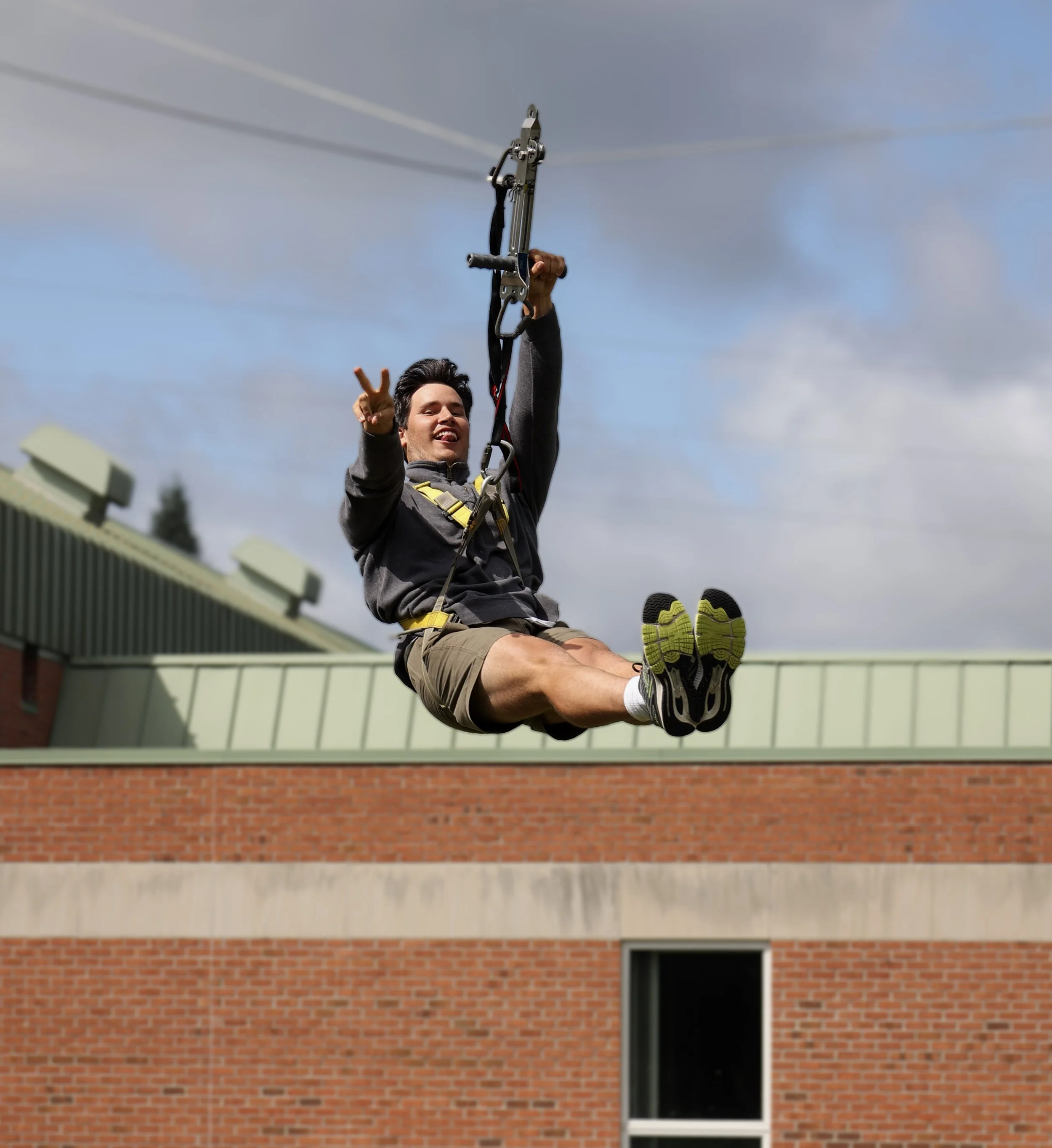 A person smiling while zip-lining in front of a brick building under a cloudy sky.