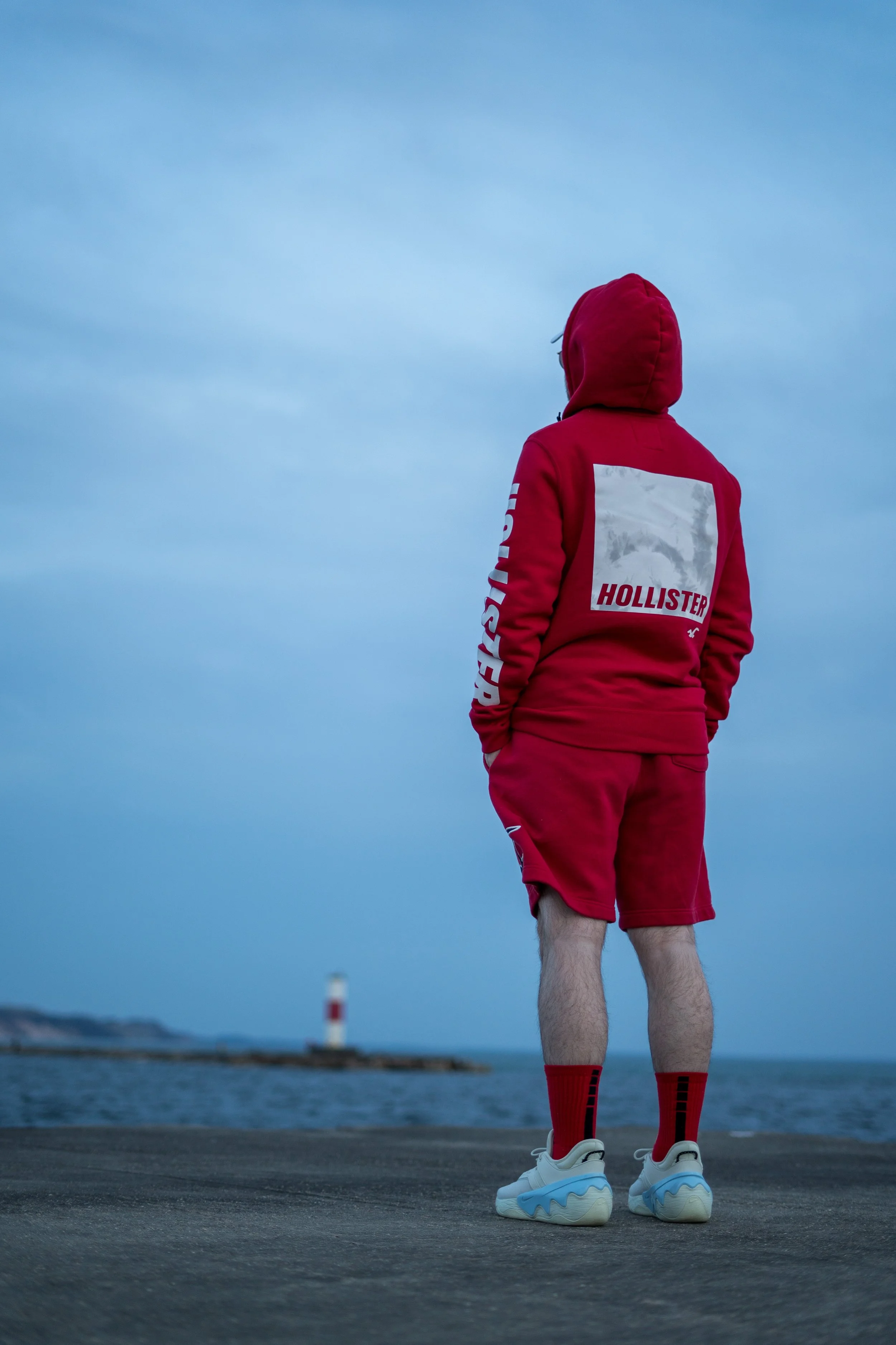Person wearing red Hollister hoodie and shorts standing by the sea with a lighthouse in the distance.