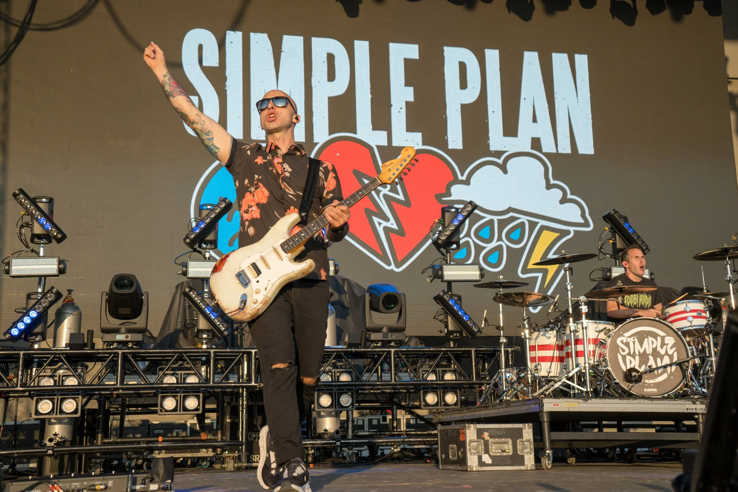 Band performing on stage with "Simple Plan" backdrop featuring heart and cloud graphics; guitarist holding white guitar and drummer on drums.