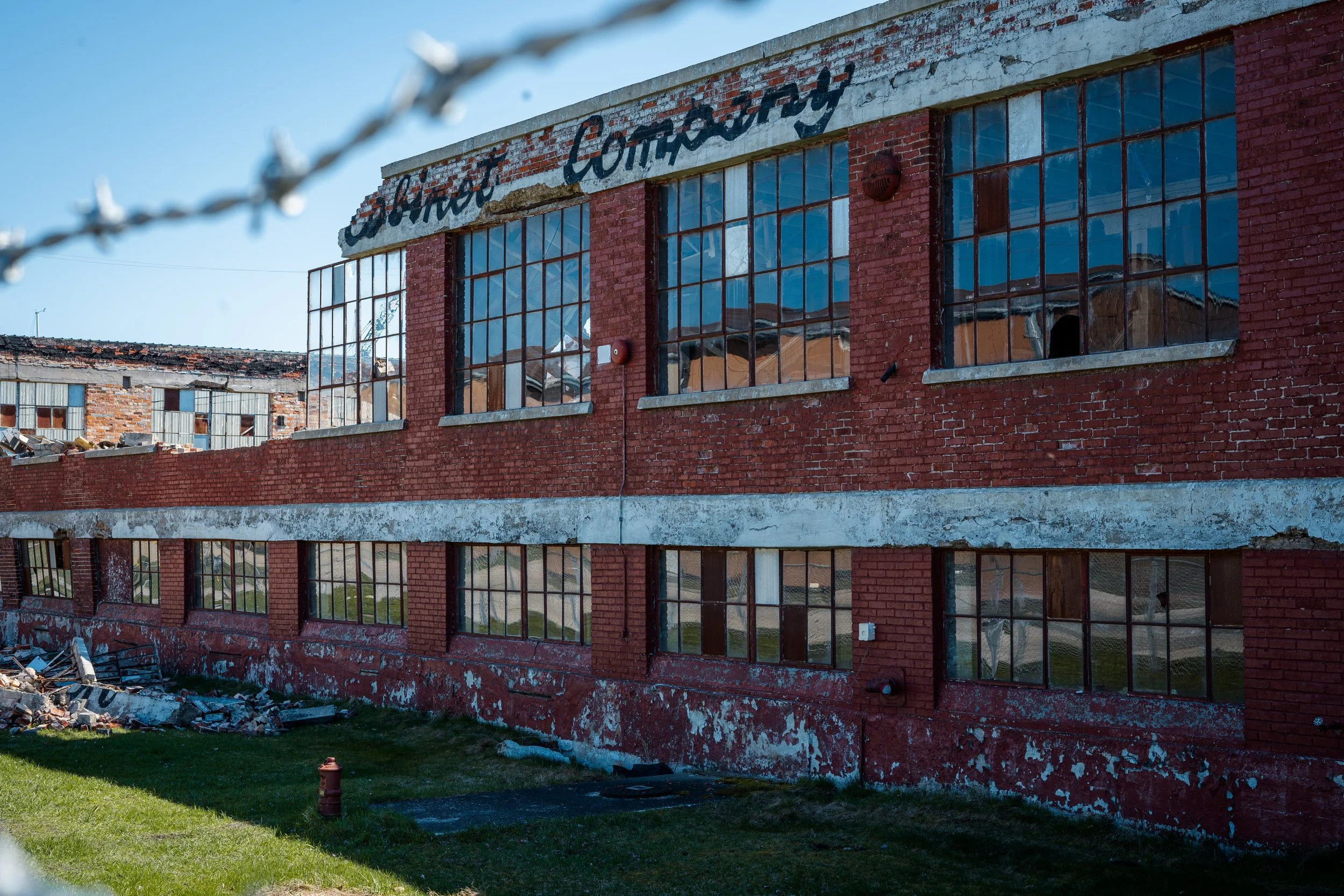 Abandoned red brick building with broken windows, barbed wire foreground, and peeling paint.