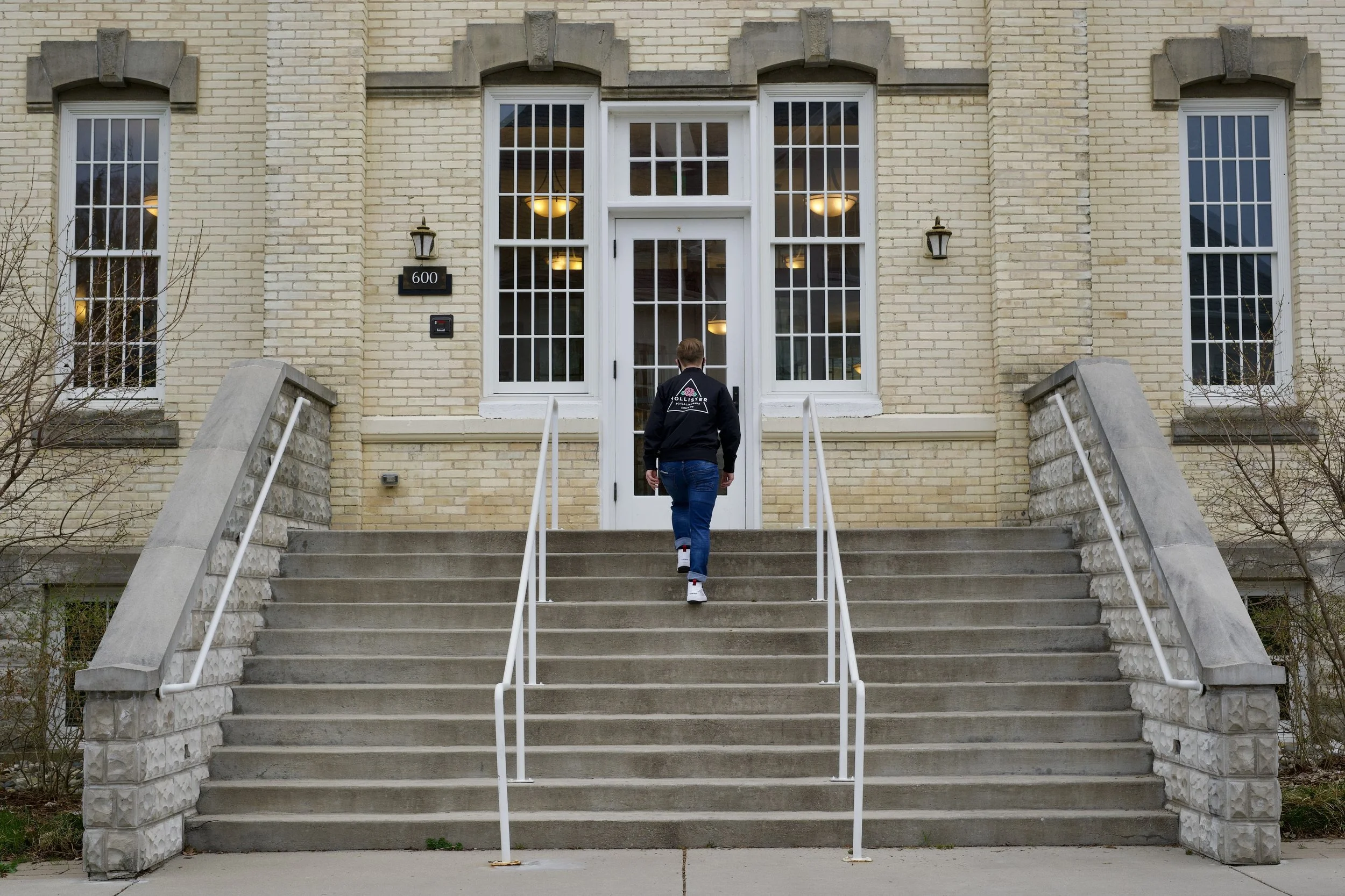 Person walking up steps to a brick building with large windows and white railings.