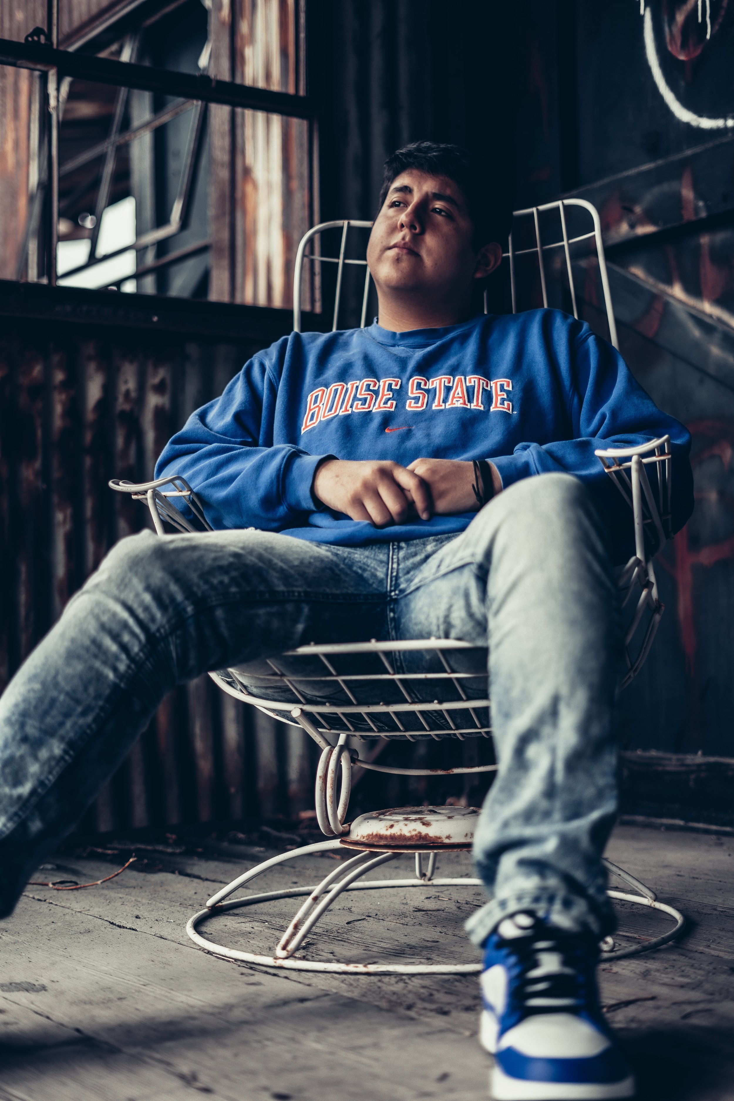 Young person sitting in a metal chair, wearing a blue "Boise State" sweatshirt and jeans in an industrial setting.