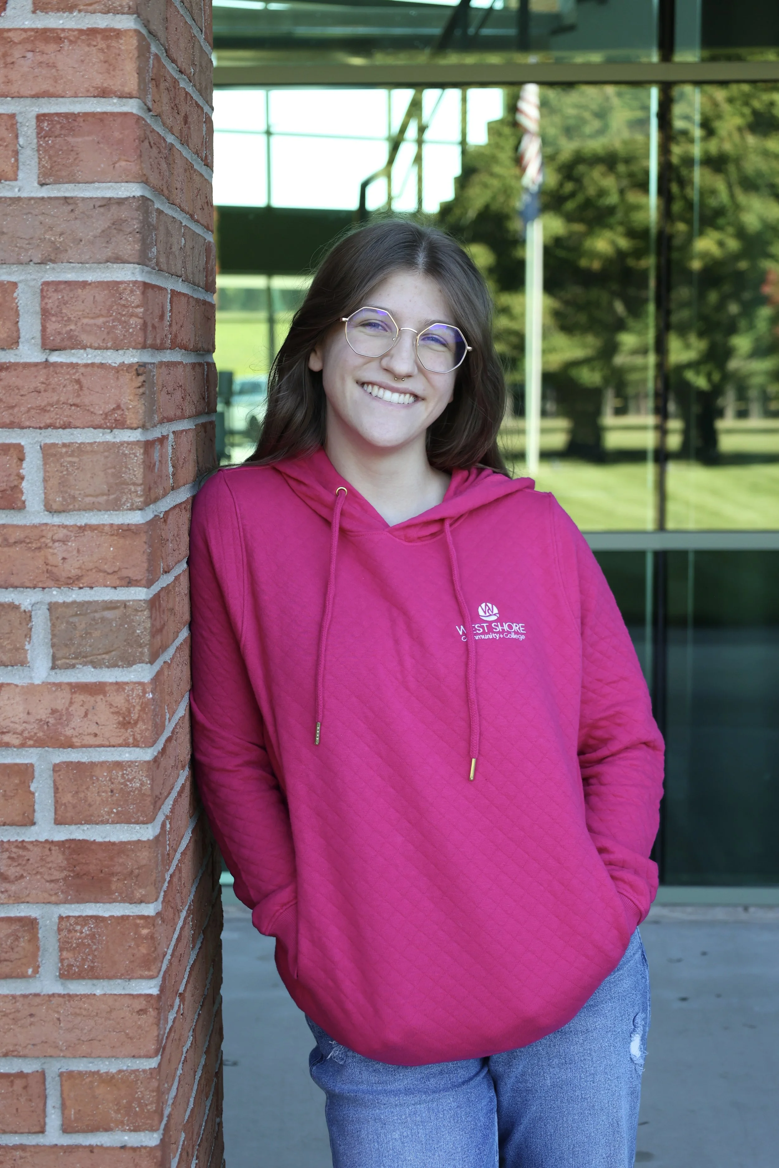 Person in a pink hoodie leaning against a brick wall, smiling.