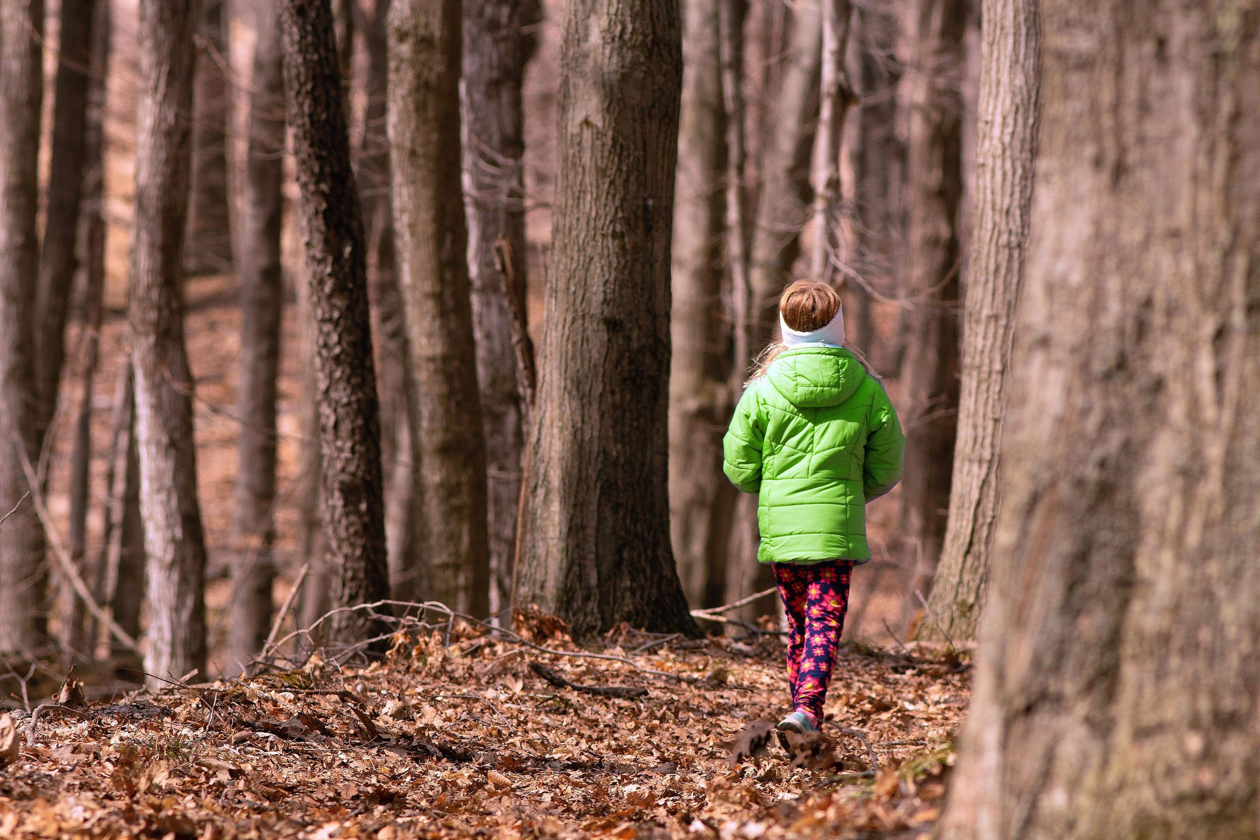 Child walking in a forest wearing a green jacket and colorful pants.