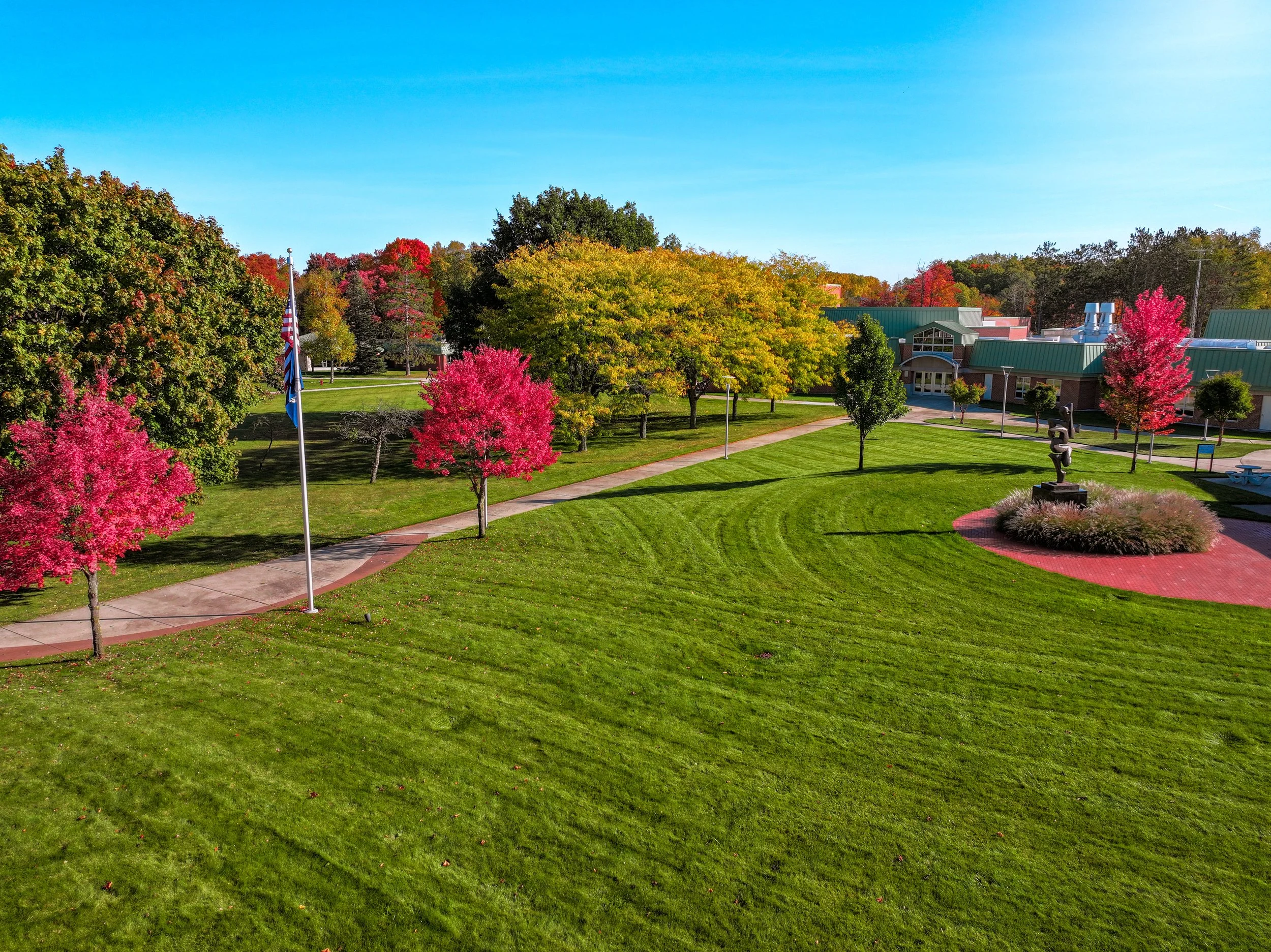 A scenic campus with green lawns, trees in autumn colors, and a pathway. There's a flagpole with an American flag, a small sculpture on a red brick circle, and a building with a green roof in the background, under a clear blue sky.
