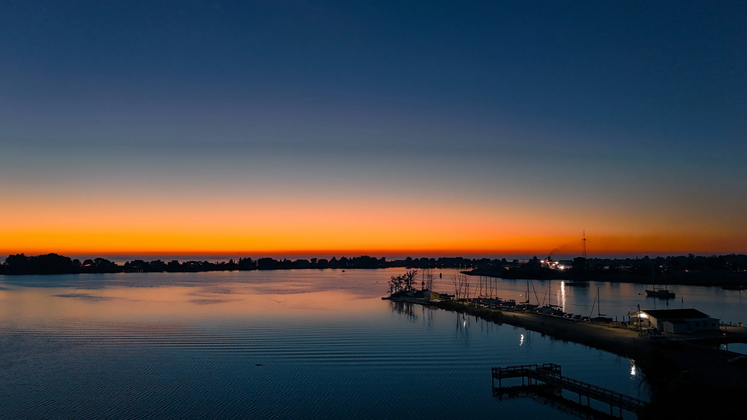 Sunset over a calm harbor with silhouetted trees and boats, orange and blue sky.