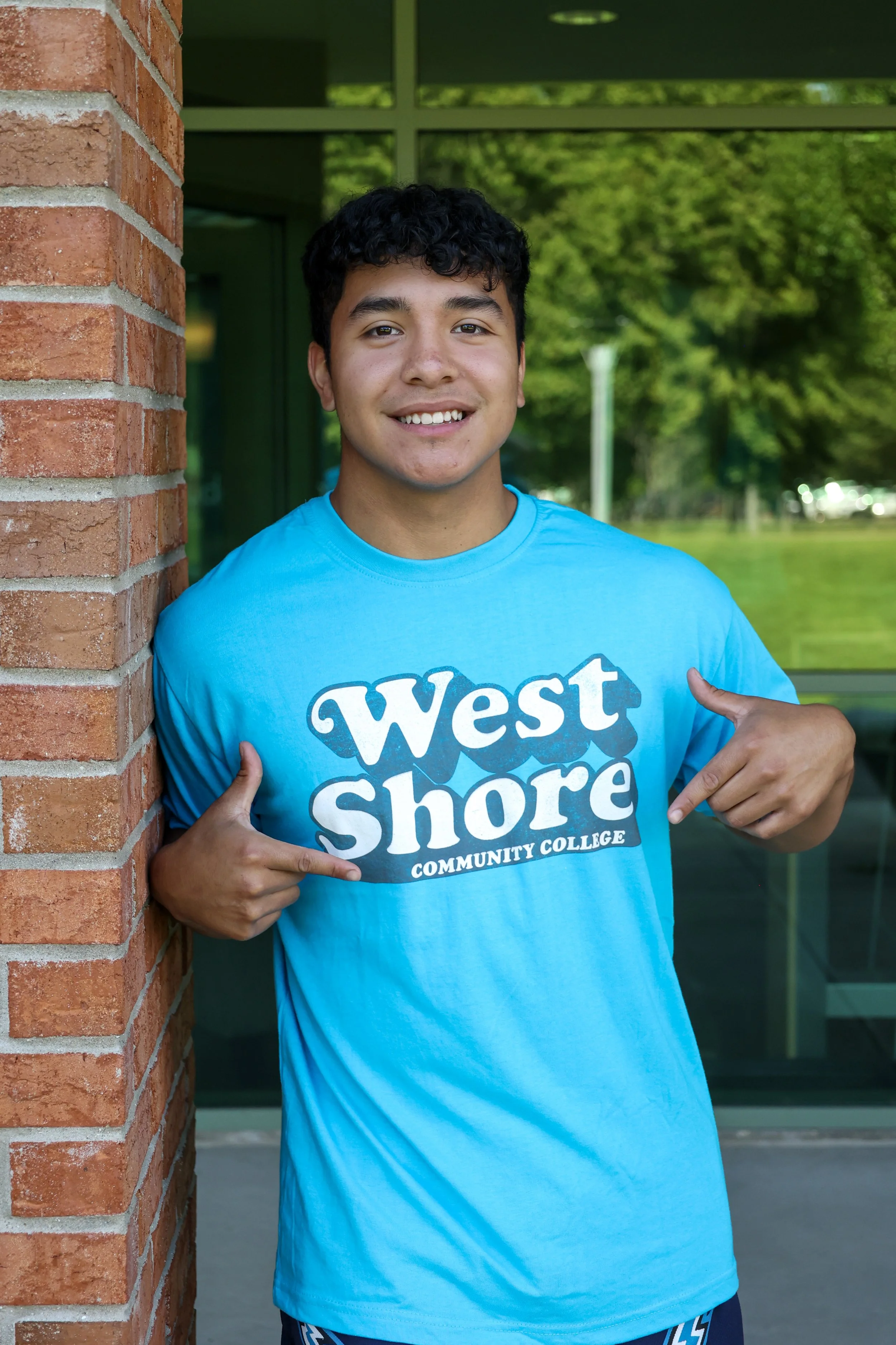 A person smiling, wearing a blue West Shore Community College t-shirt, standing outdoors next to a brick wall.