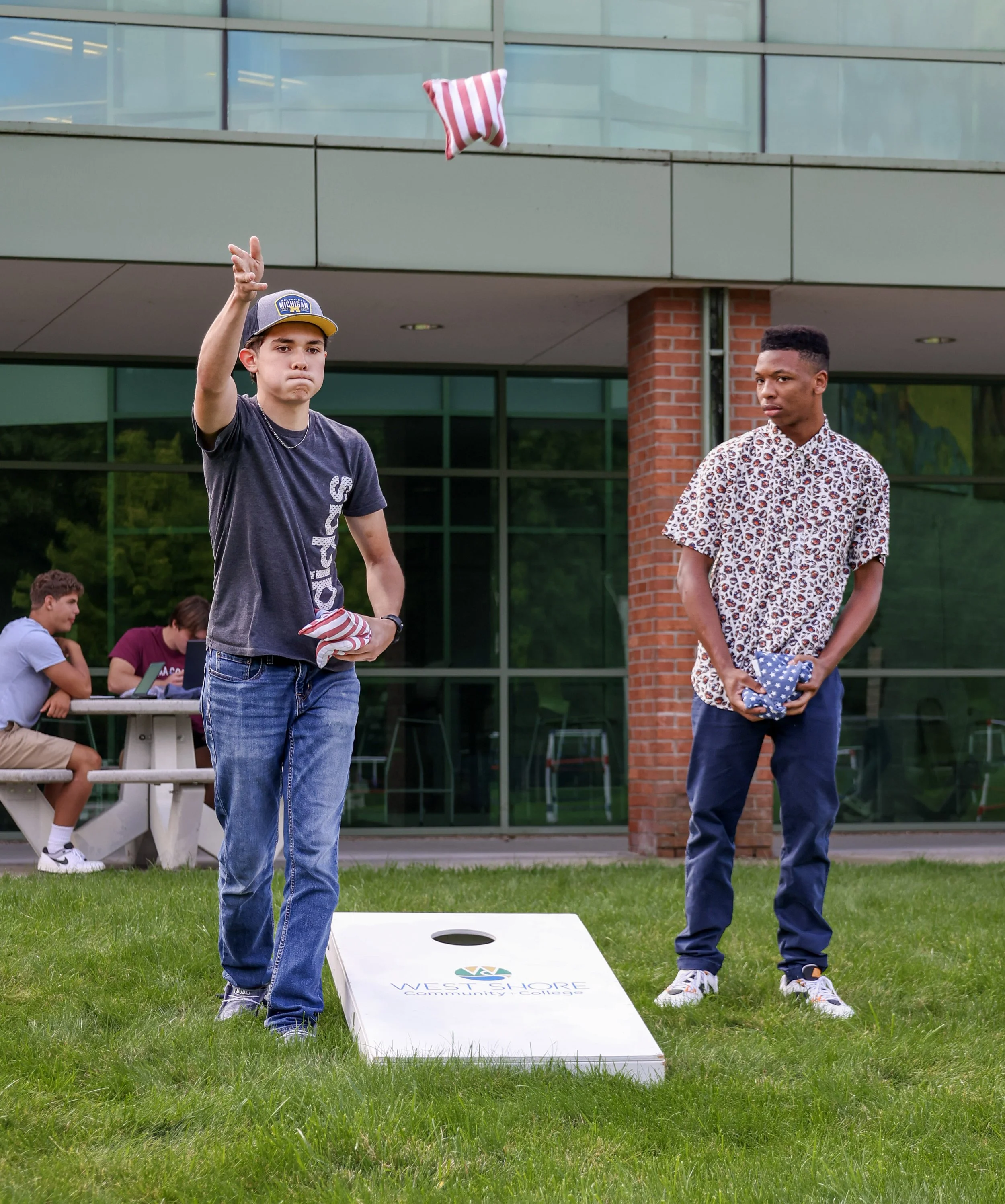 Two young men playing cornhole on a grassy lawn with a cornhole board marked "Westshore Community College." One man is tossing a bean bag while the other holds bean bags, standing in front of a building with glass windows.
