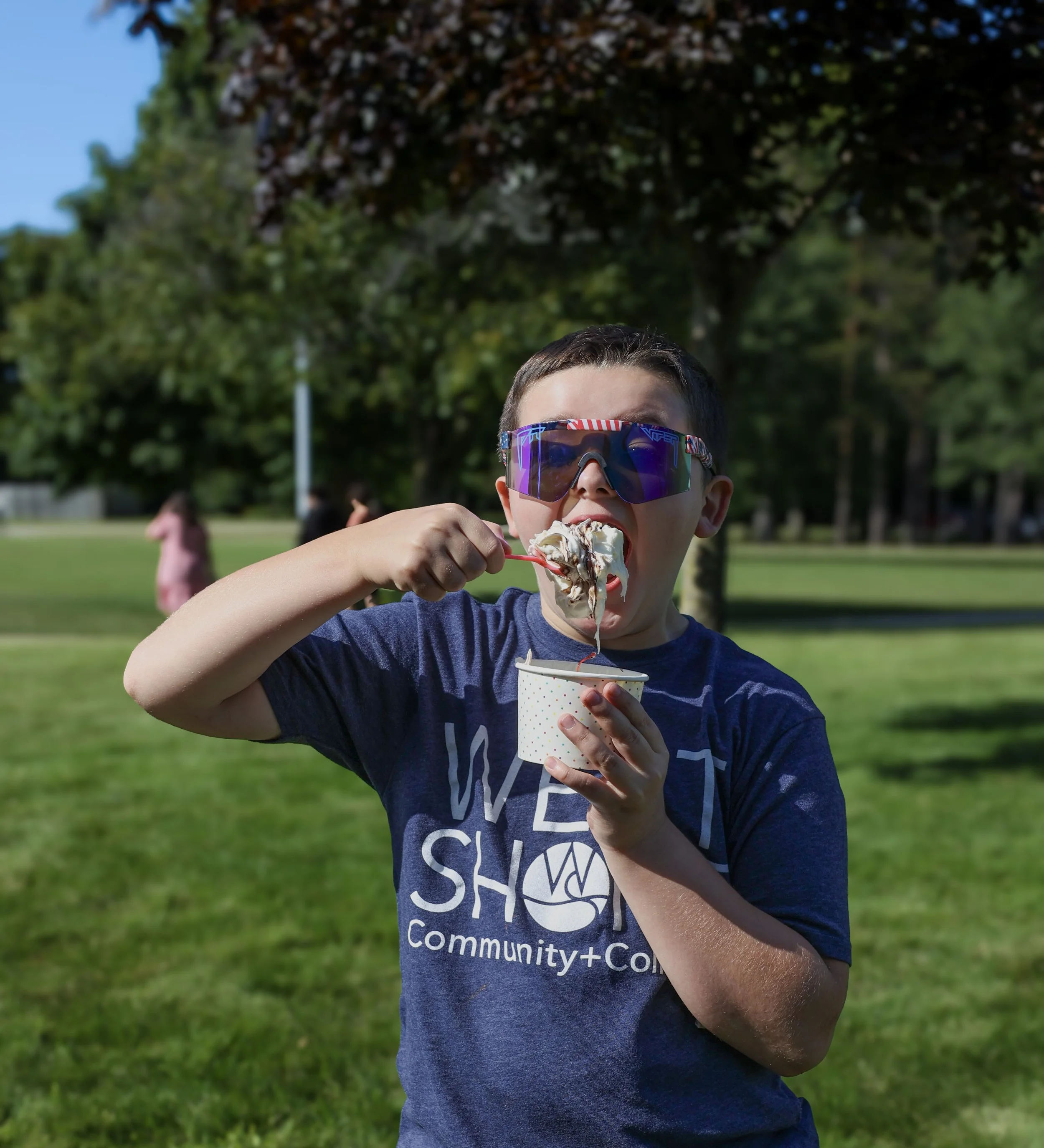 Child eating ice cream in a park, wearing sunglasses and a "West Shore Community College" t-shirt.