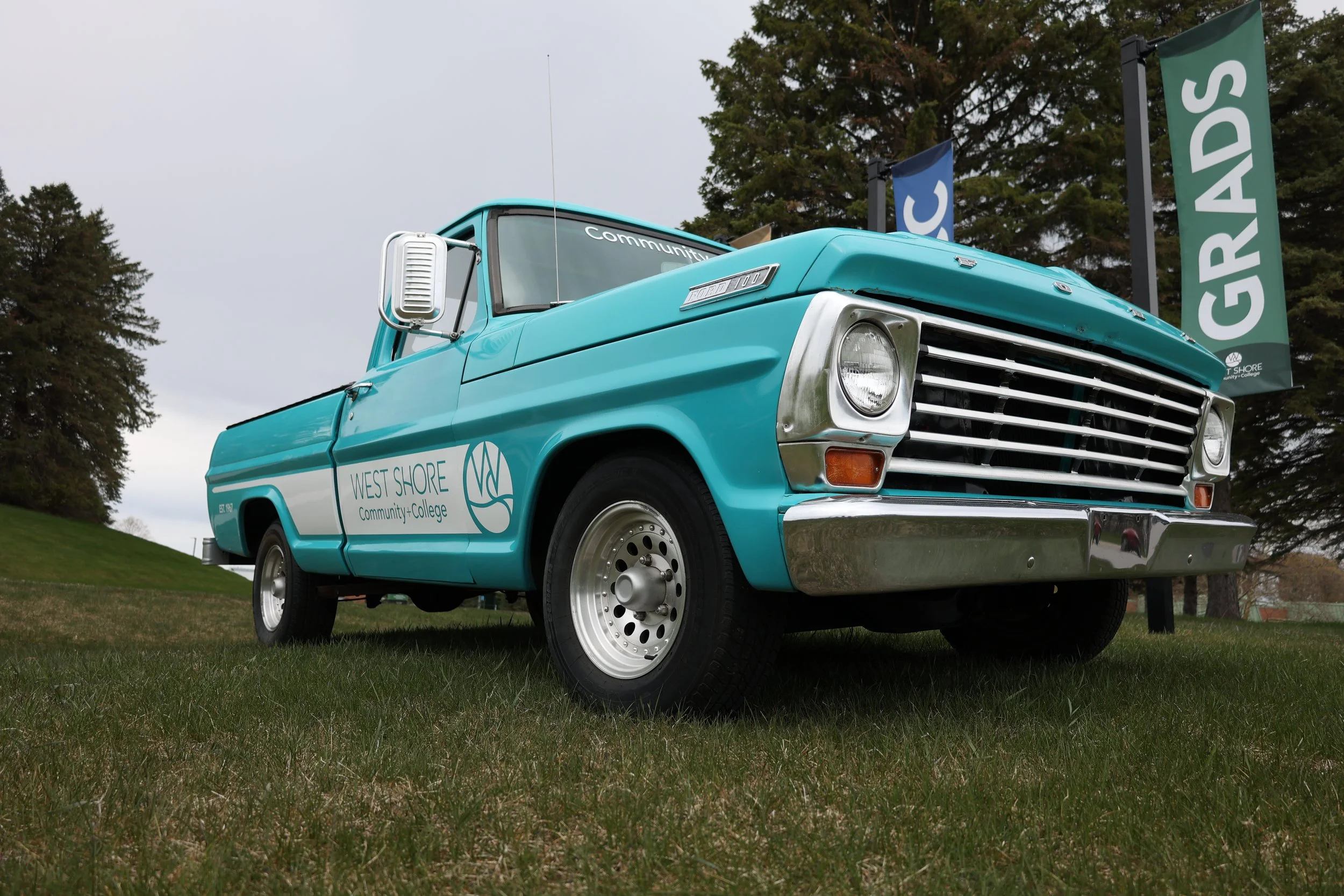 Classic turquoise Ford pickup truck with "West Shore Community College" logo, parked on grass with graduation banners in the background.