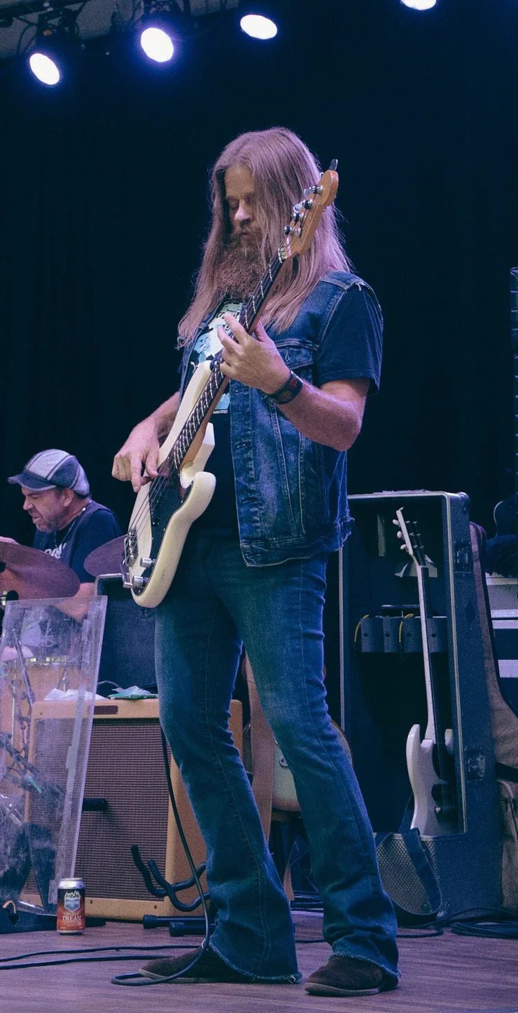 Musician playing a bass guitar on stage, wearing denim and casual attire, with a drummer playing in the background.