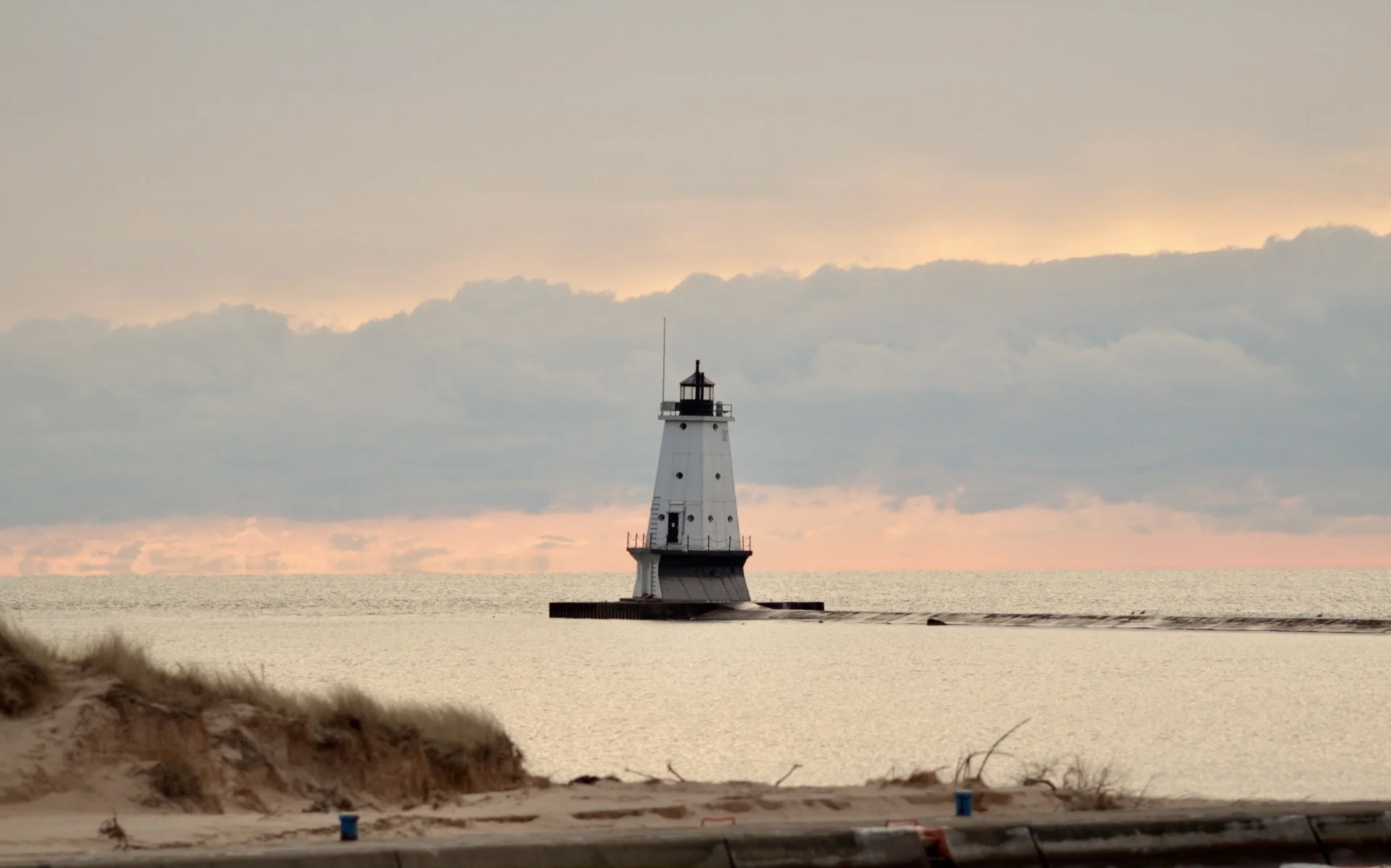 Lighthouse on a pier extending into a calm body of water with a sandy beach in the foreground, under a cloudy sky at sunrise or sunset.