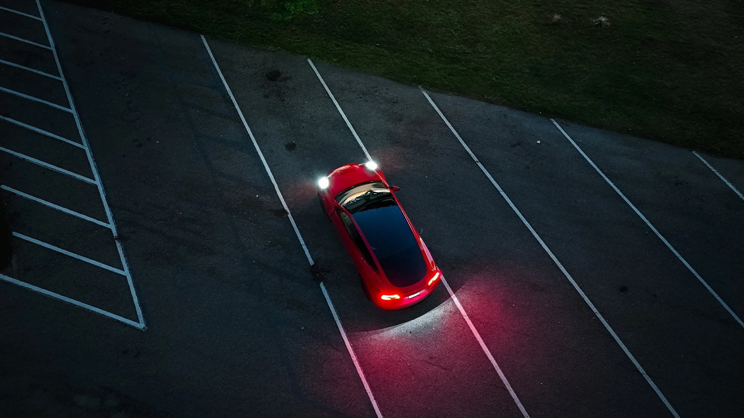Aerial view of a red car with headlights on in an empty parking lot at night.