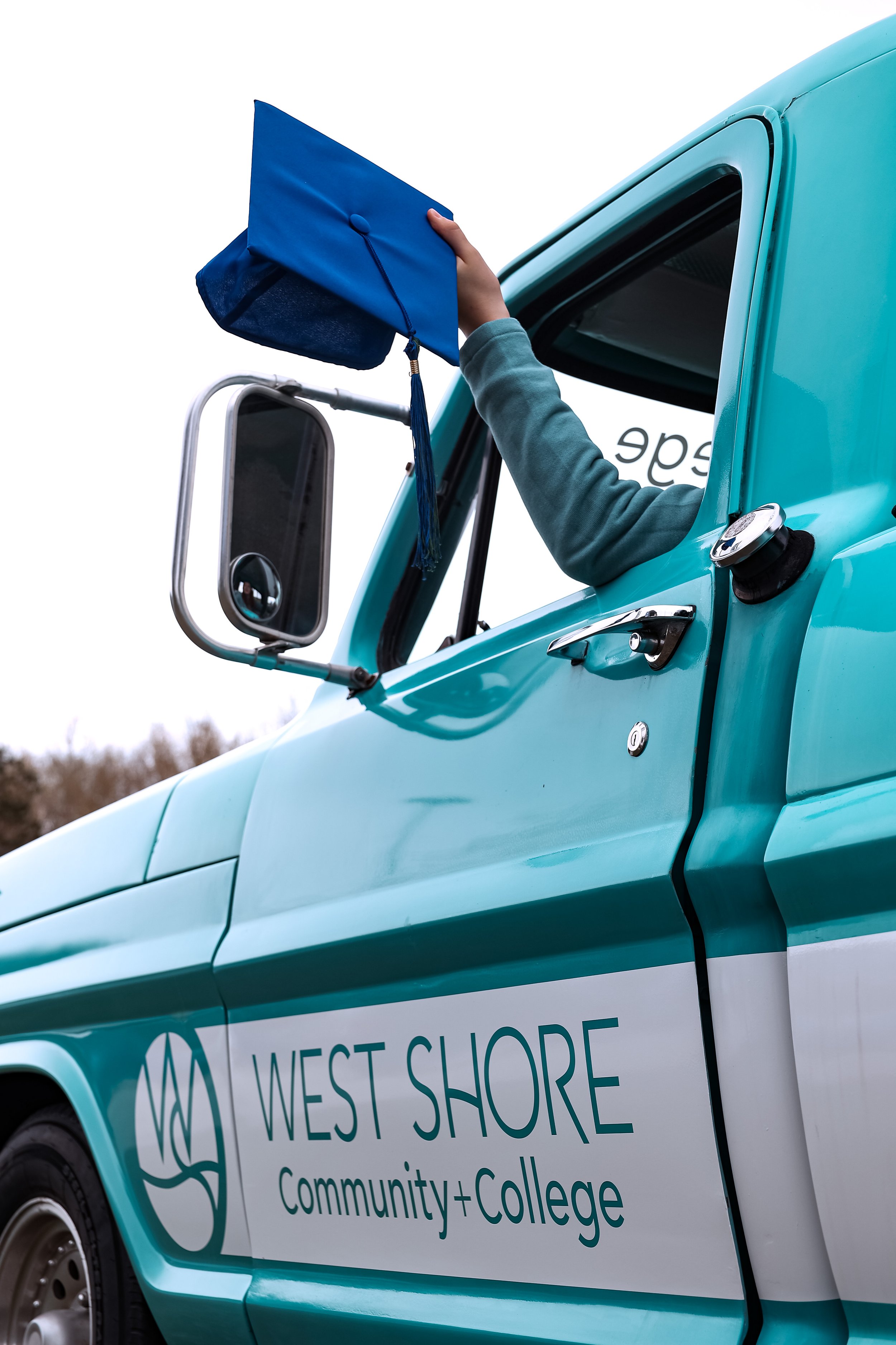 Person in a turquoise truck displaying a blue graduation cap with "West Shore Community College" sign on the door.