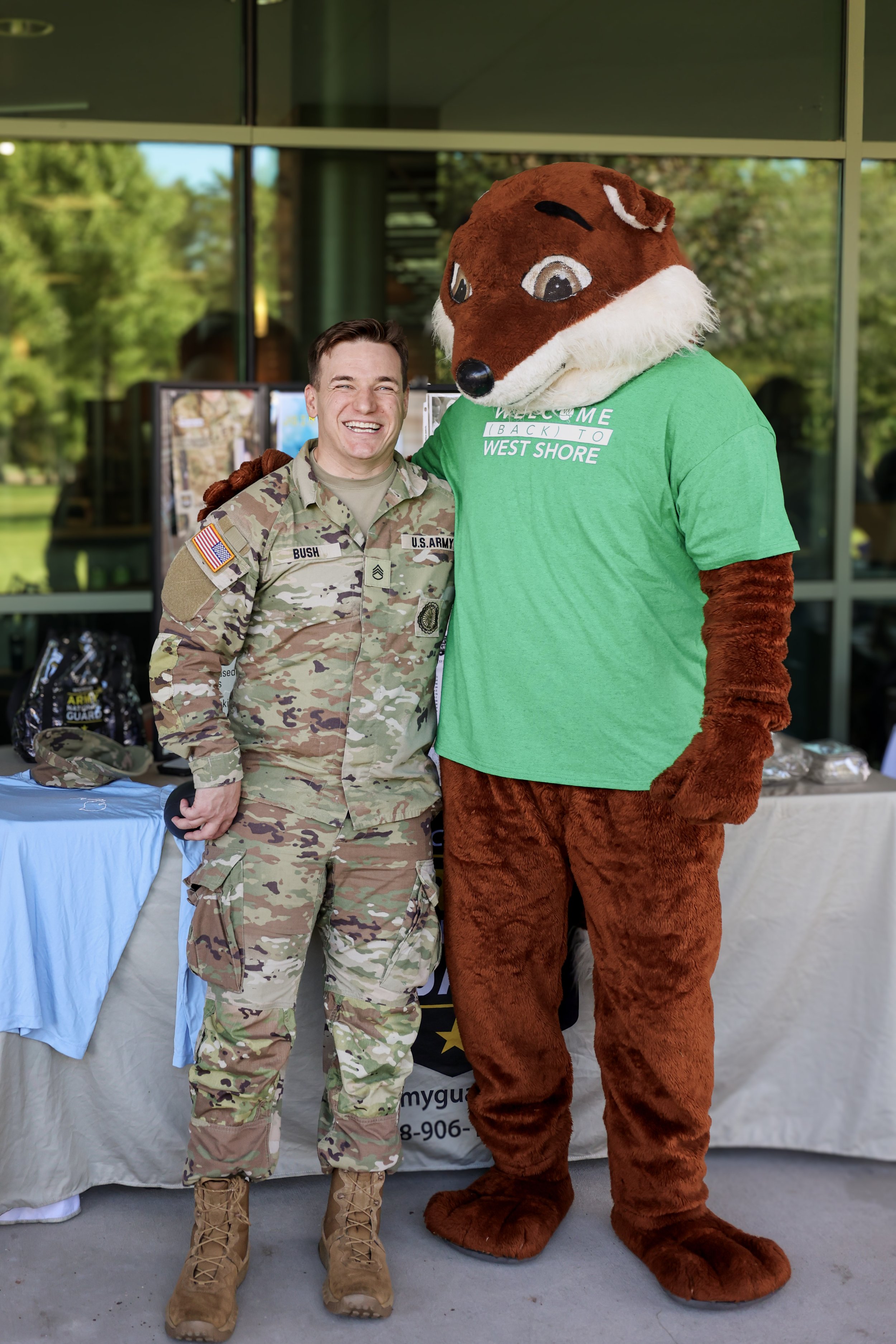 A uniformed military personnel smiling beside a person dressed in a fox mascot costume wearing a green "Welcome Back to West Shore" T-shirt, standing in front of a table with promotional items.