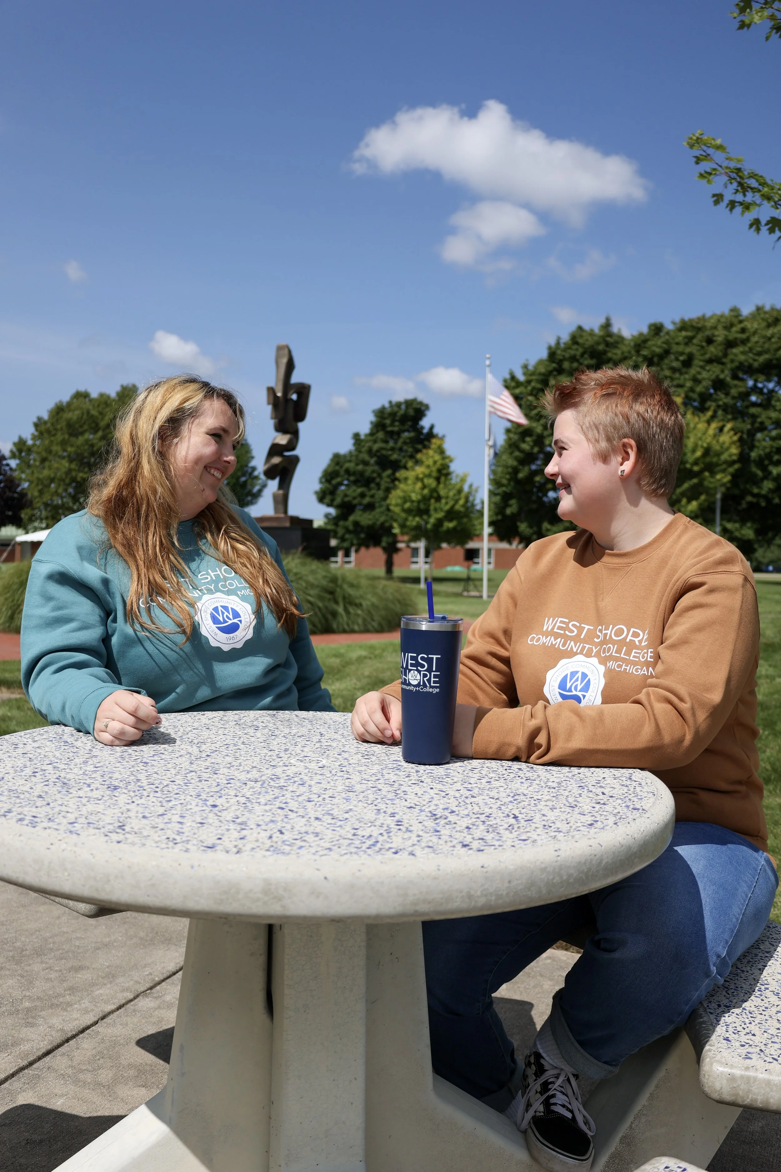 Two people sitting at a concrete outdoor table, wearing "West Shore Community College" sweatshirts. They are smiling and conversing. An American flag and trees are in the background on a sunny day.