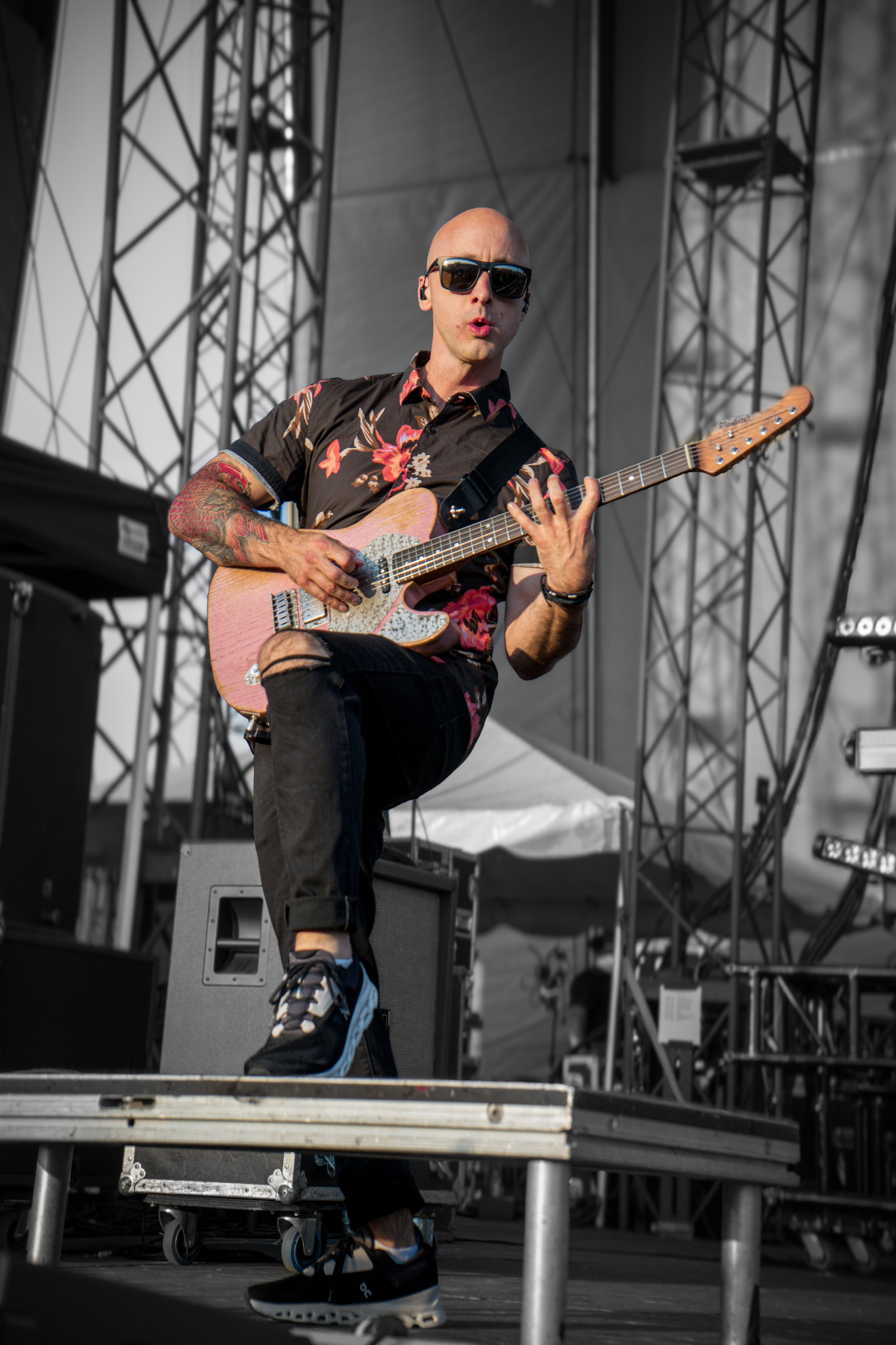 Guitarist performing on stage, wearing sunglasses and a floral shirt, holding an electric guitar with stage equipment in the background.