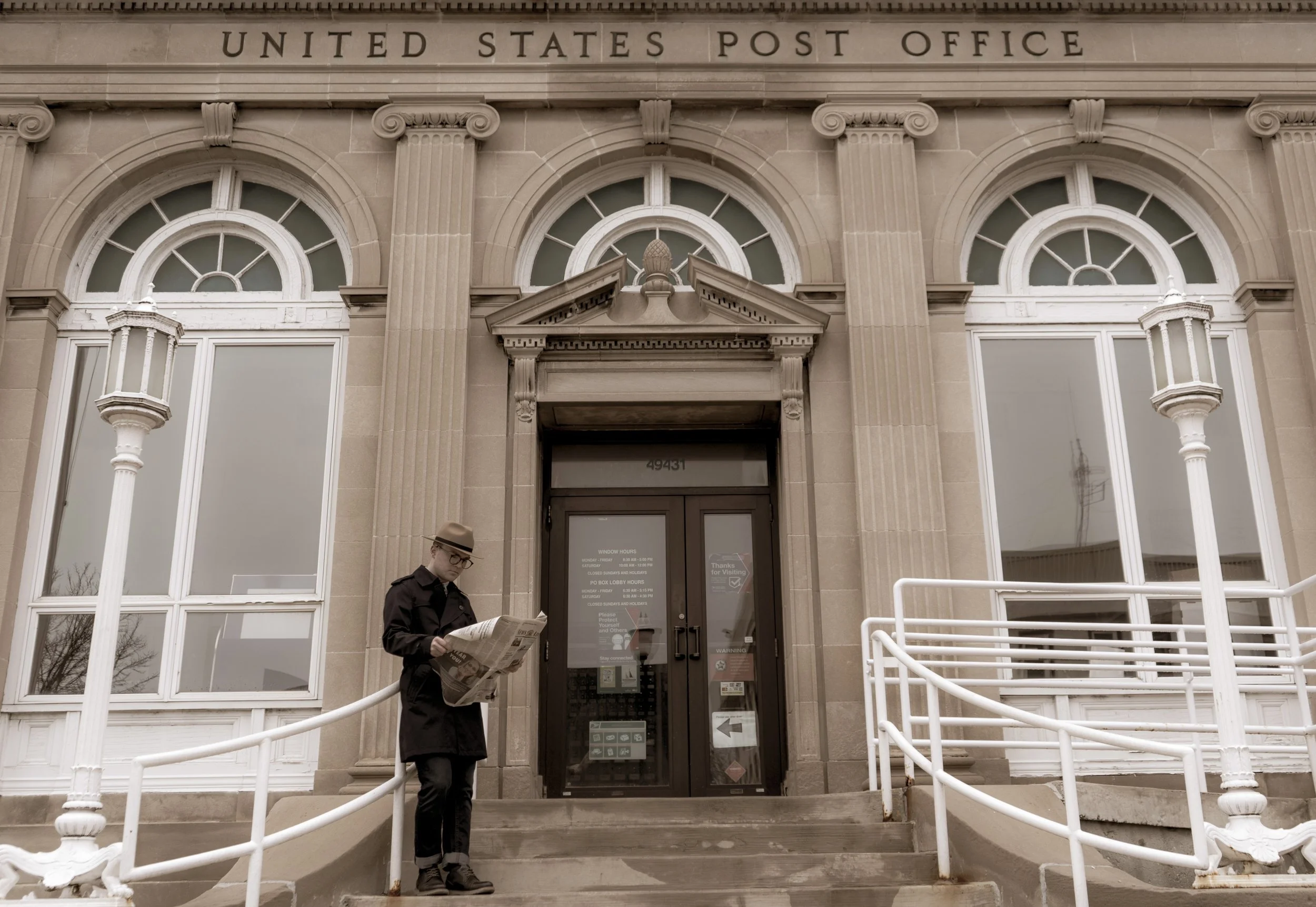Man reading a newspaper on the steps of a historic United States Post Office building with columns and arched windows.