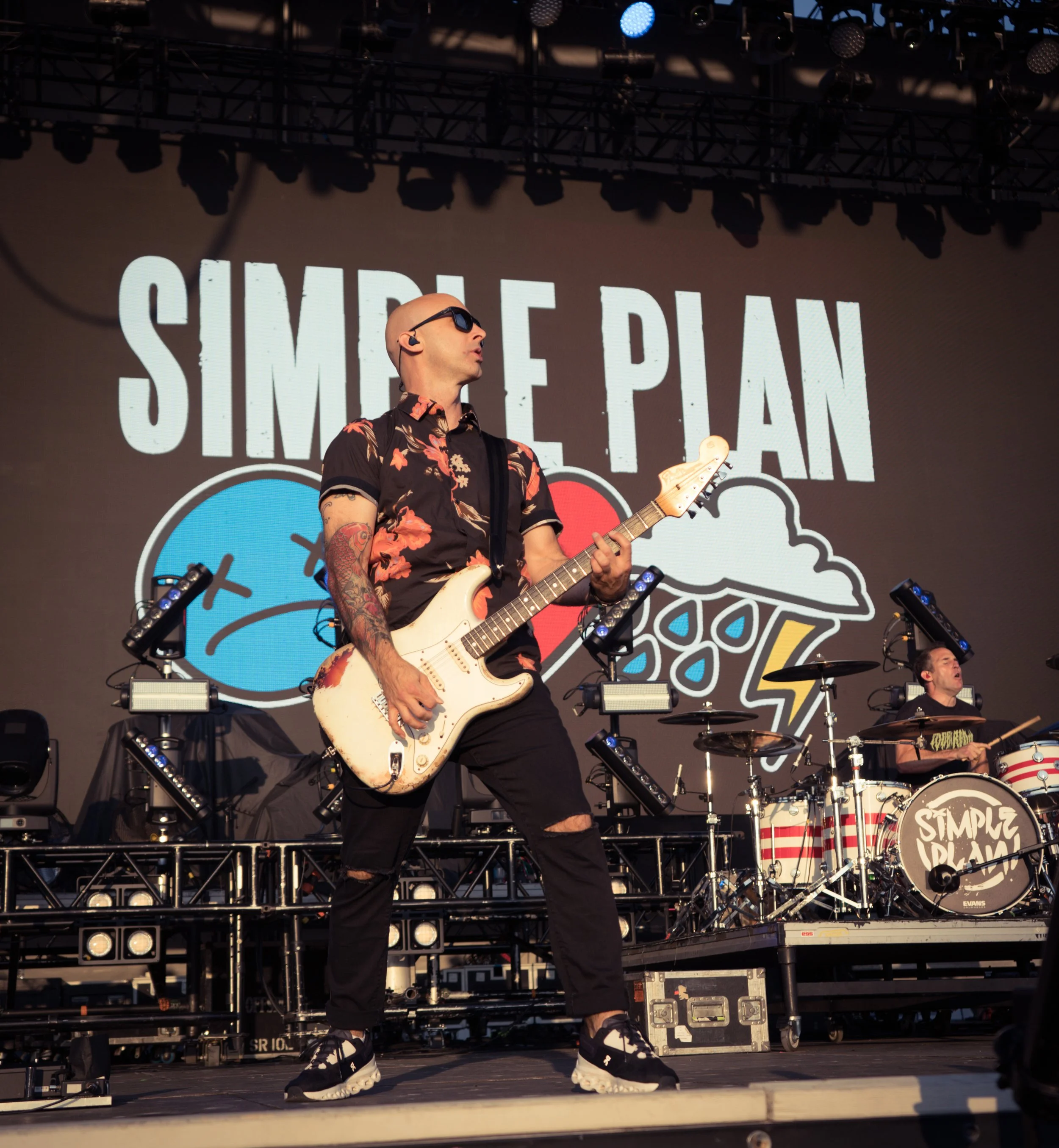 A guitarist on stage performing with a drummer in the background, with the "Simple Plan" logo displayed on the screen behind them.