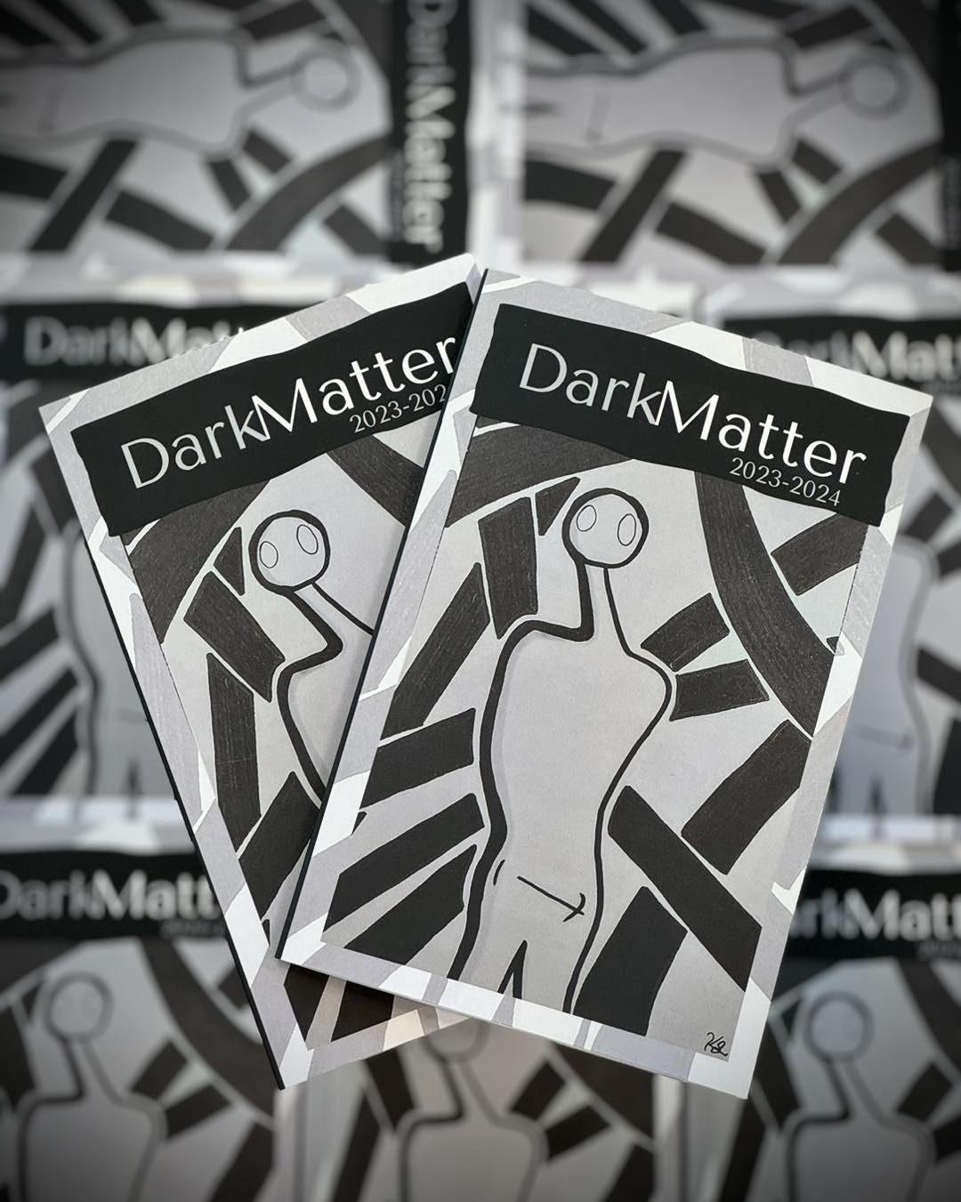 Two copies of "Dark Matter 2023-2024" magazine with abstract black and white cover art featuring a stylized humanoid figure and geometric patterns.