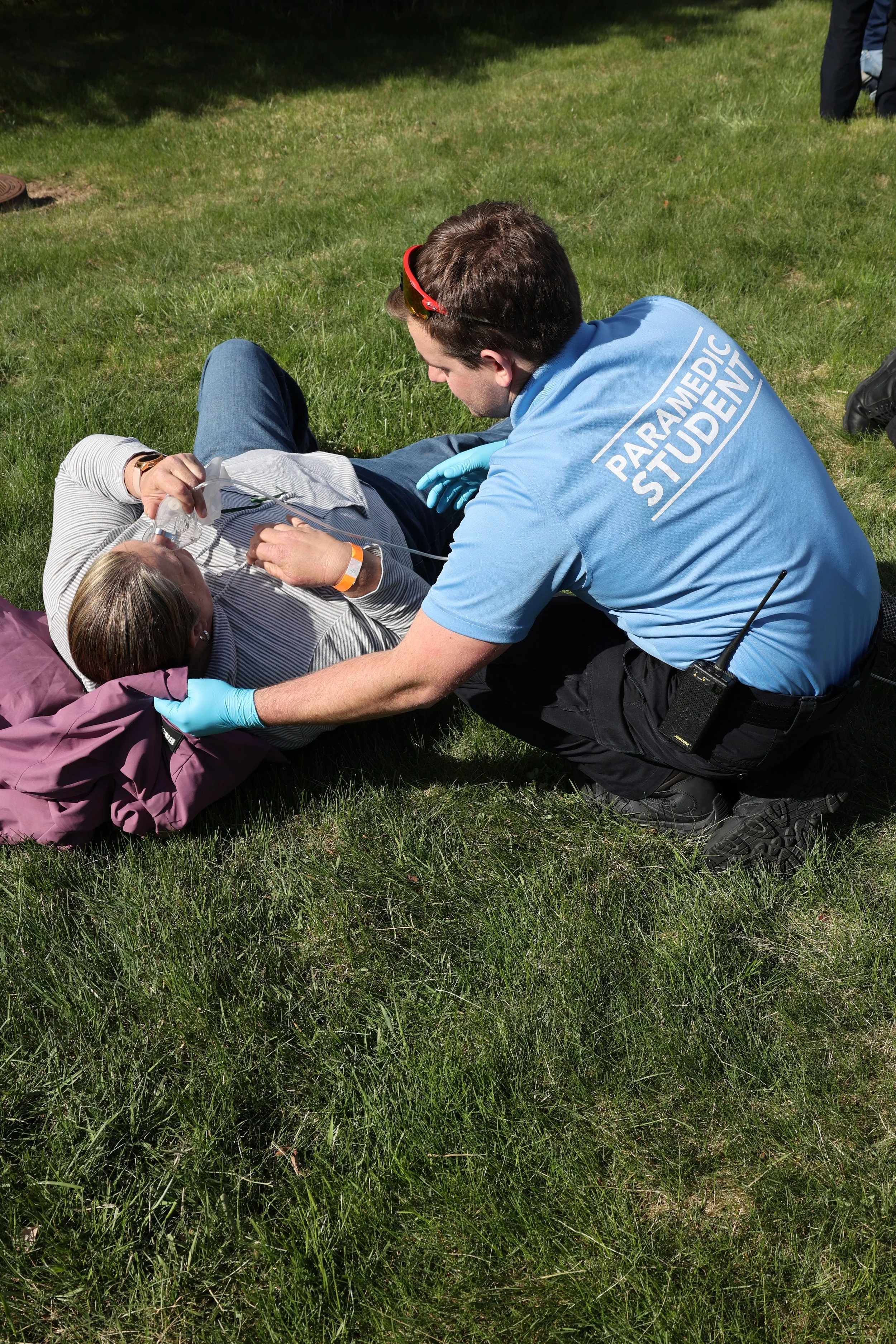 A paramedic student assisting a person lying on grass, providing oxygen through a face mask, with gloved hands.