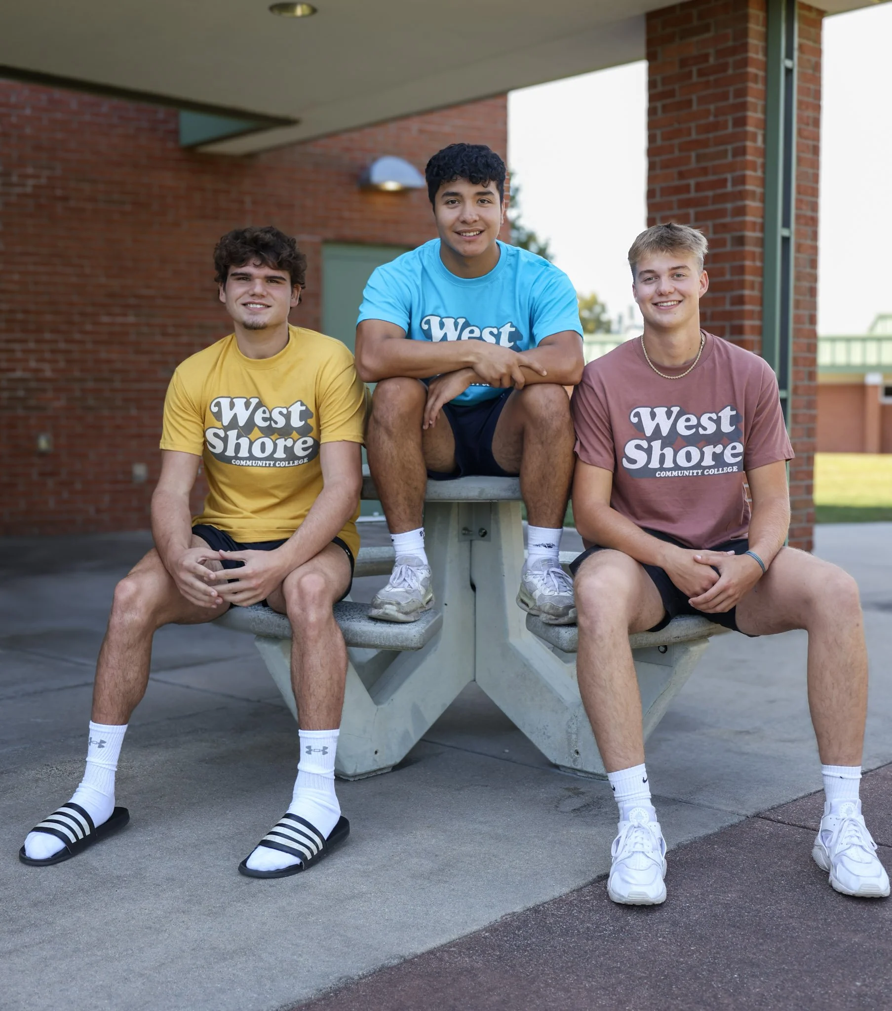 Three young men sitting outdoors wearing "West Shore Community College" t-shirts in different colors, with one sitting on a table and the others on the bench. Brick wall in the background.