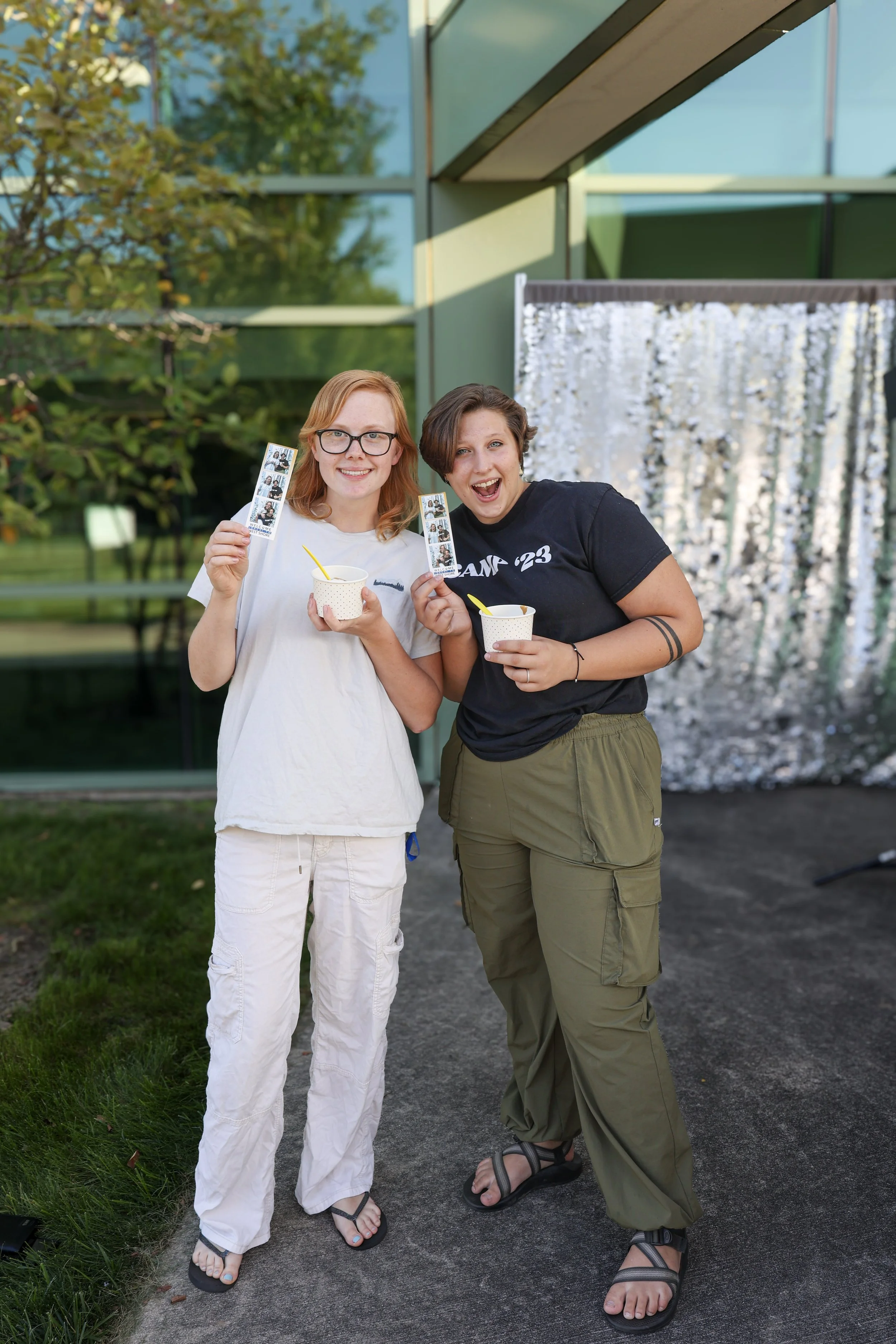 Two people smiling, holding photo booth strips and ice cream cups, standing outdoors in front of a building with a reflective backdrop.