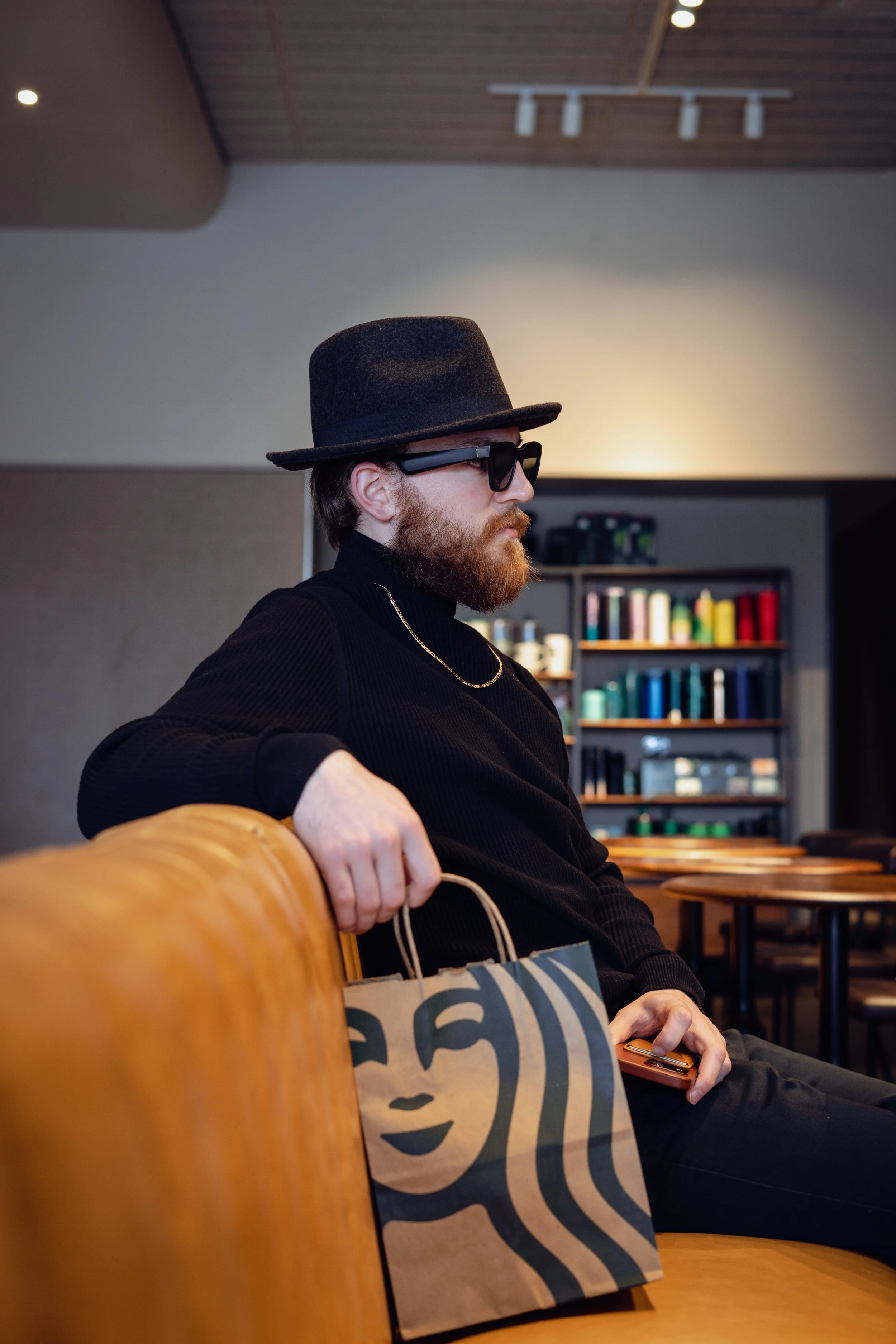 Man sitting in a cafe holding a paper bag with a logo, wearing sunglasses and a hat, with shelves and tables in the background.