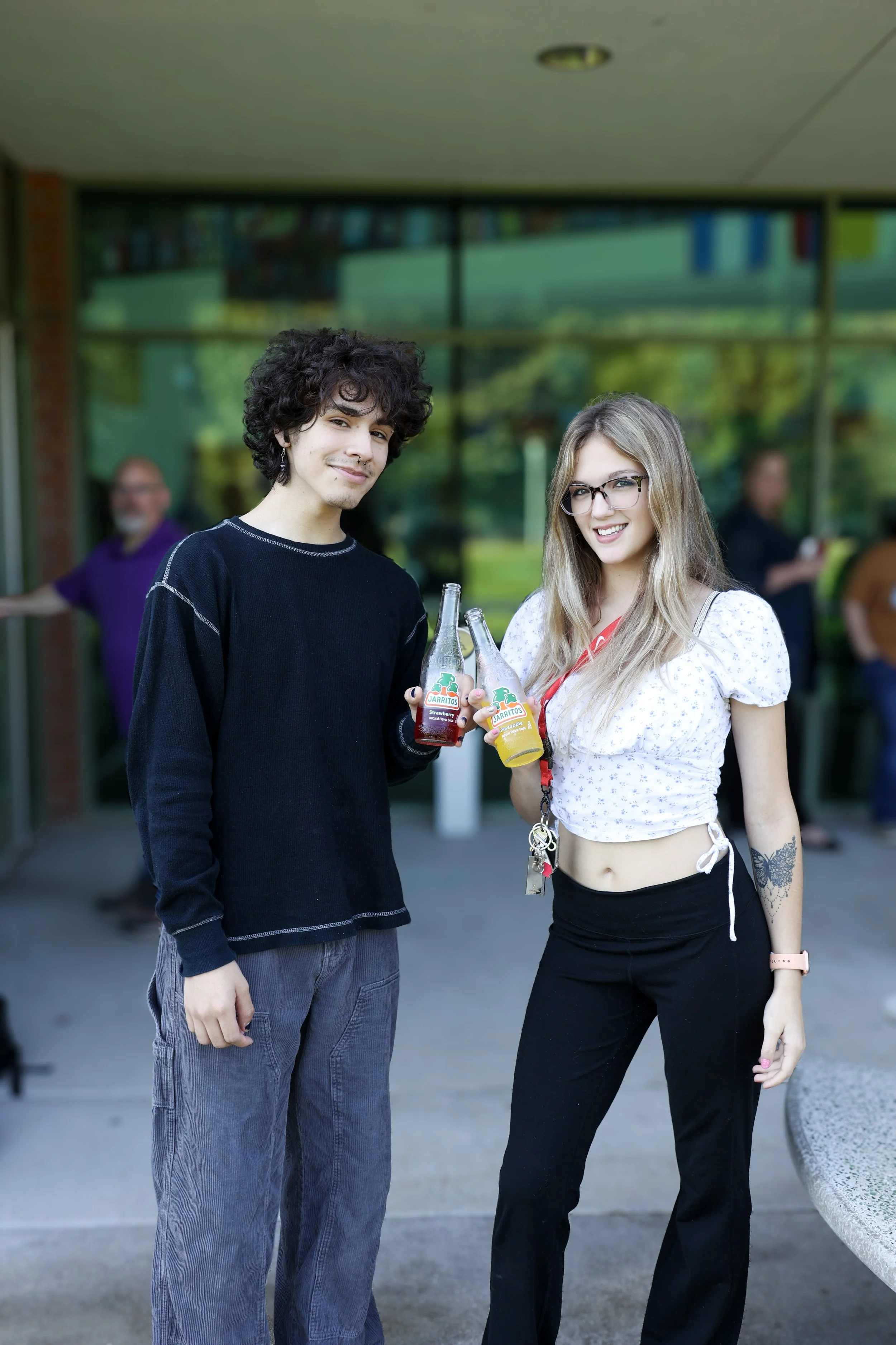 Two people standing outside holding bottles of Jarritos soda, smiling at the camera, with a building and others in the background.