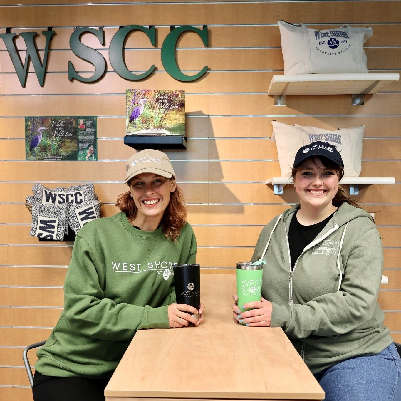 Two women in green West Shore Community College attire, smiling and holding drink containers, seated at a wooden table in front of WSCC merchandise display.