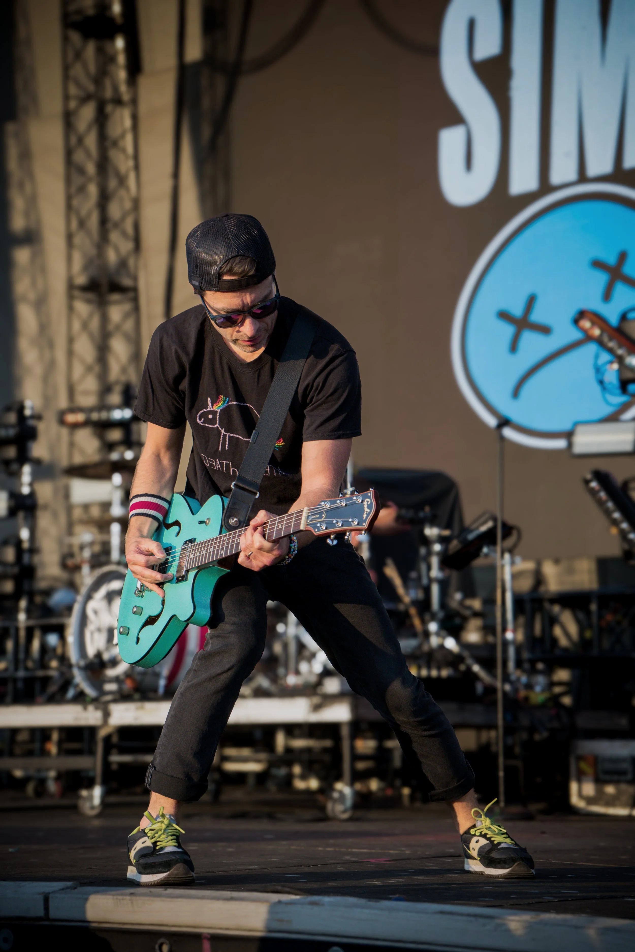 Guitarist performing on stage with teal electric guitar, wearing a black cap, sunglasses, and a black T-shirt.