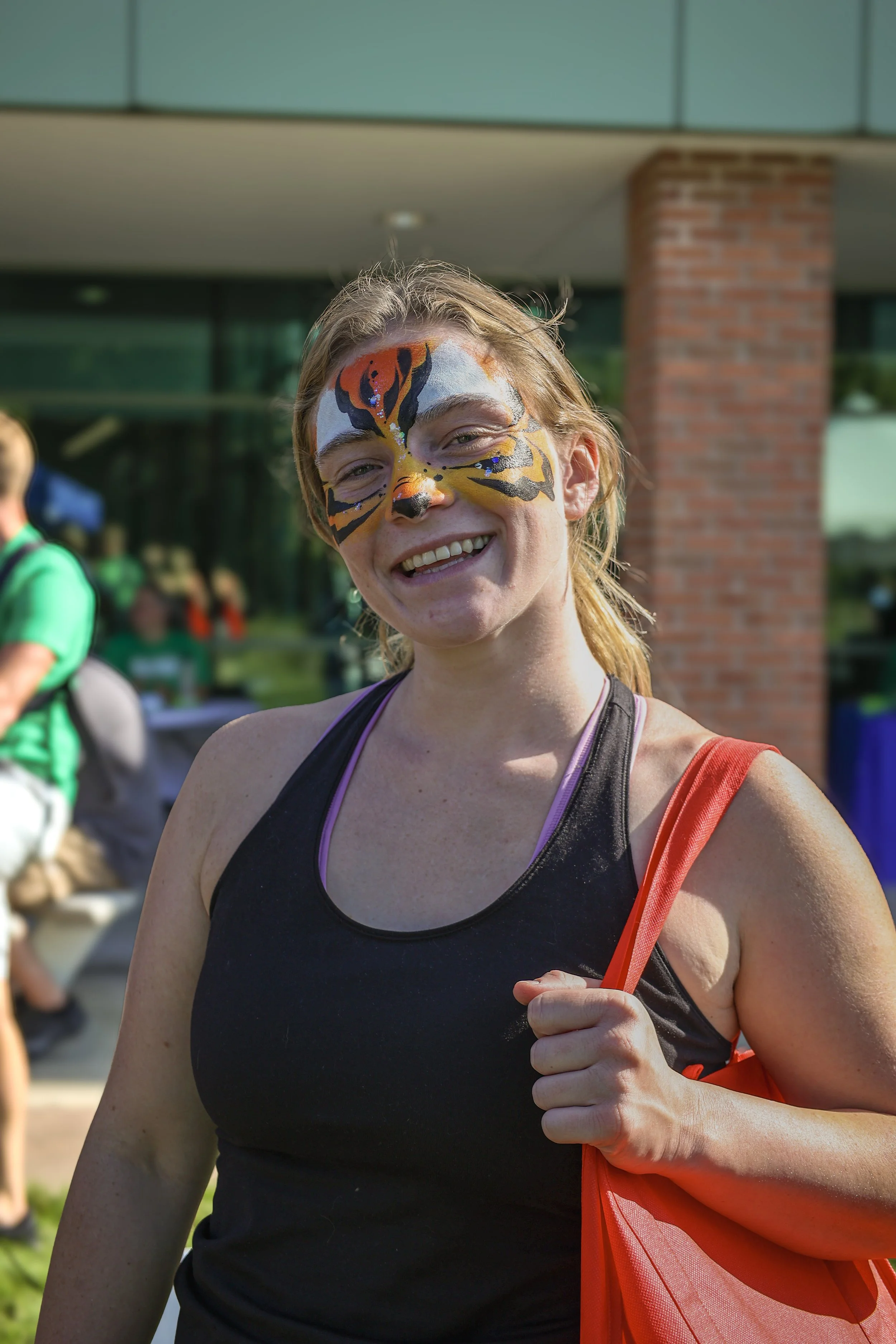 Person smiling with tiger face paint, holding an orange bag outdoors.