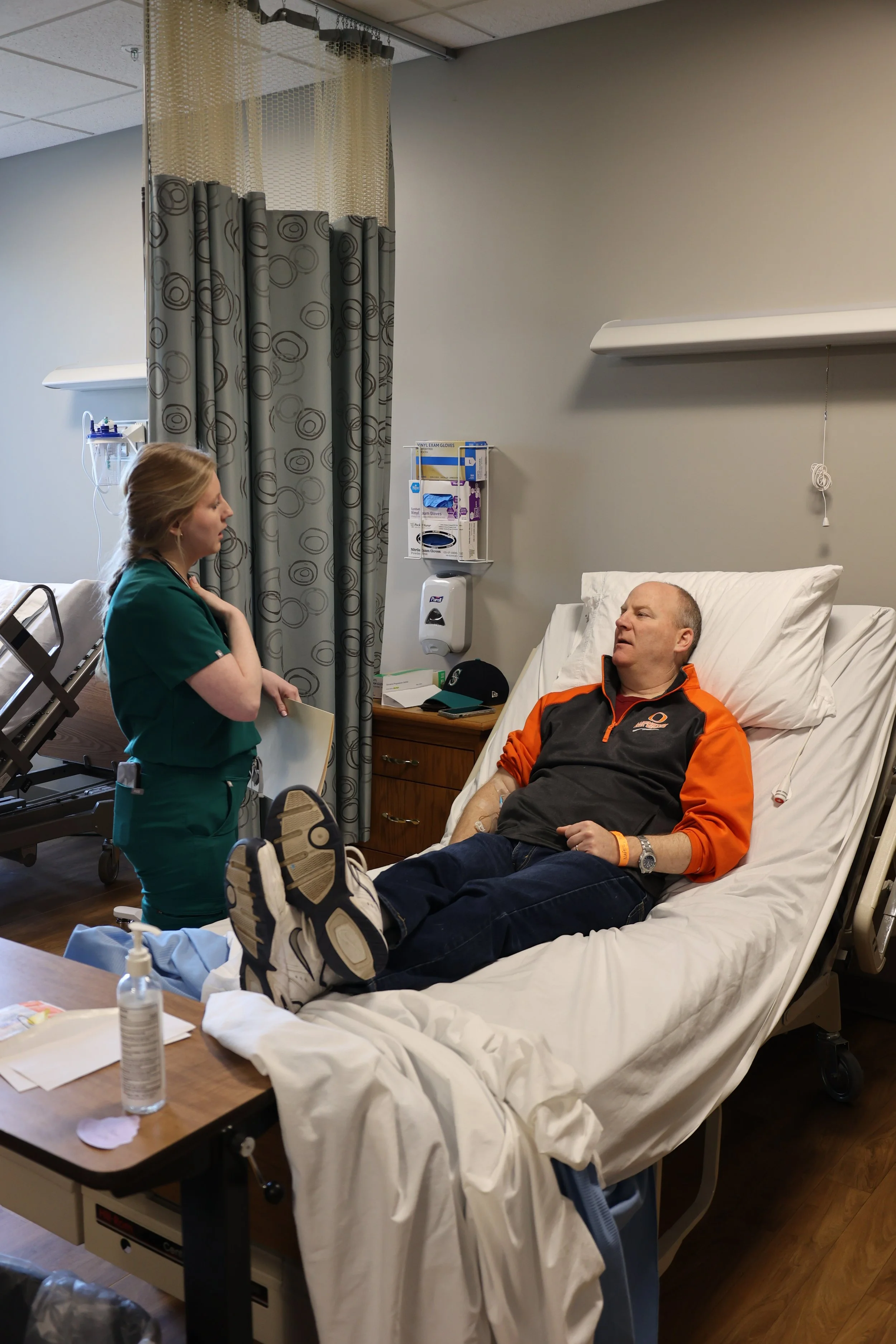 Healthcare worker in green scrubs talking to a patient in a hospital bed.