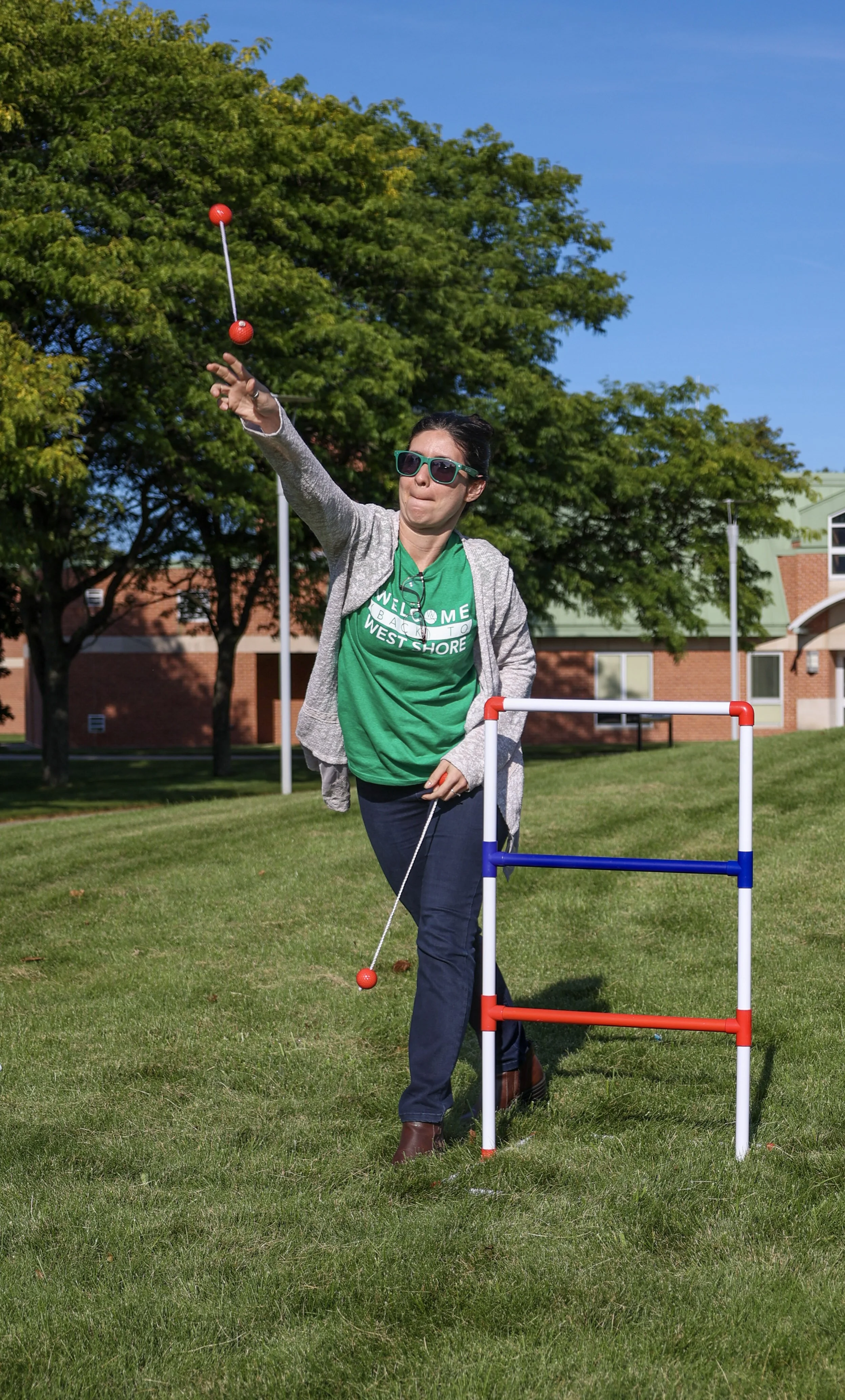 Person playing ladder toss outdoors in a grassy area on a sunny day, wearing sunglasses and a green t-shirt.