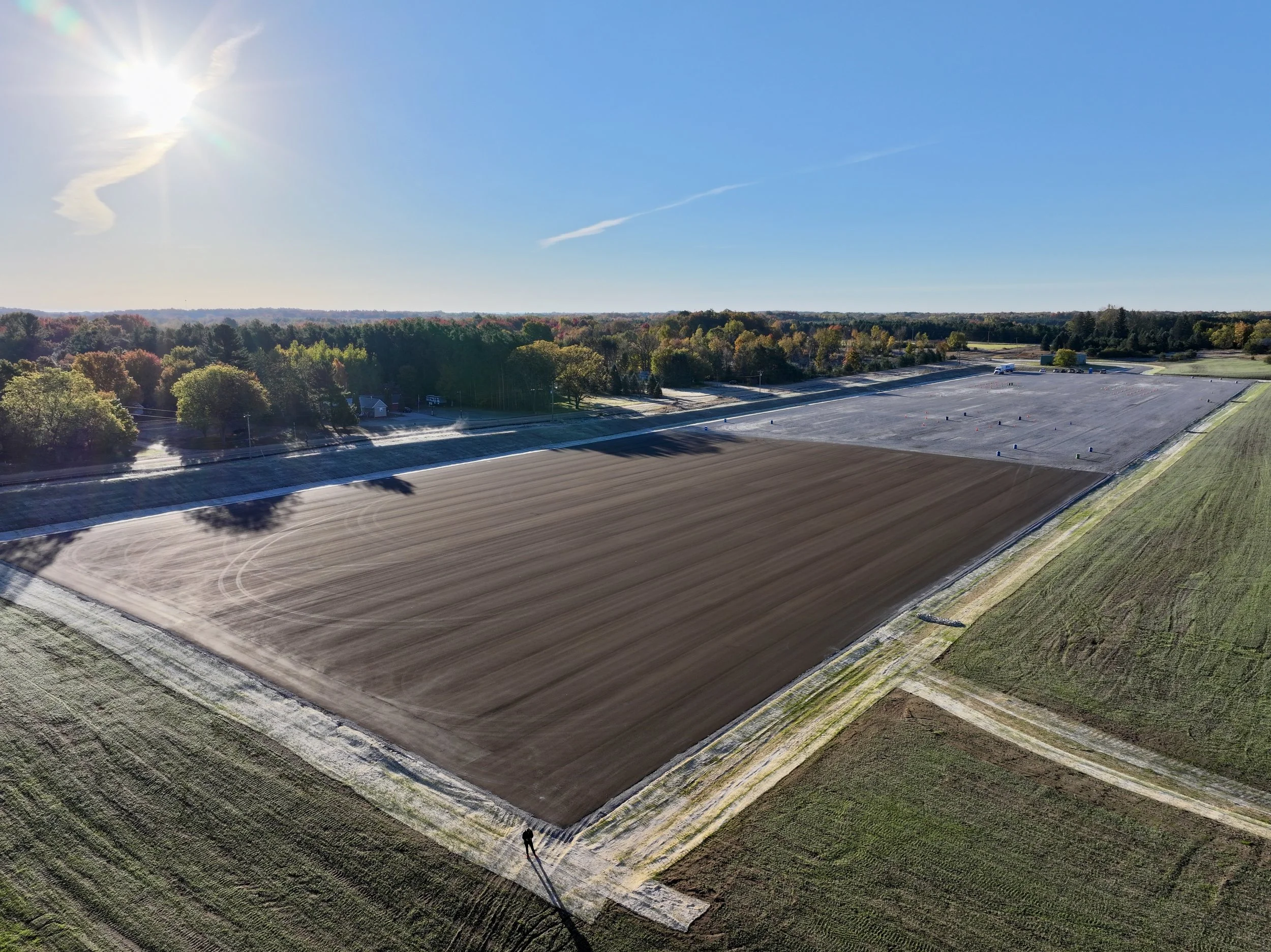 Aerial view of a large, paved open area surrounded by greenery and trees under a clear blue sky with the sun shining brightly.