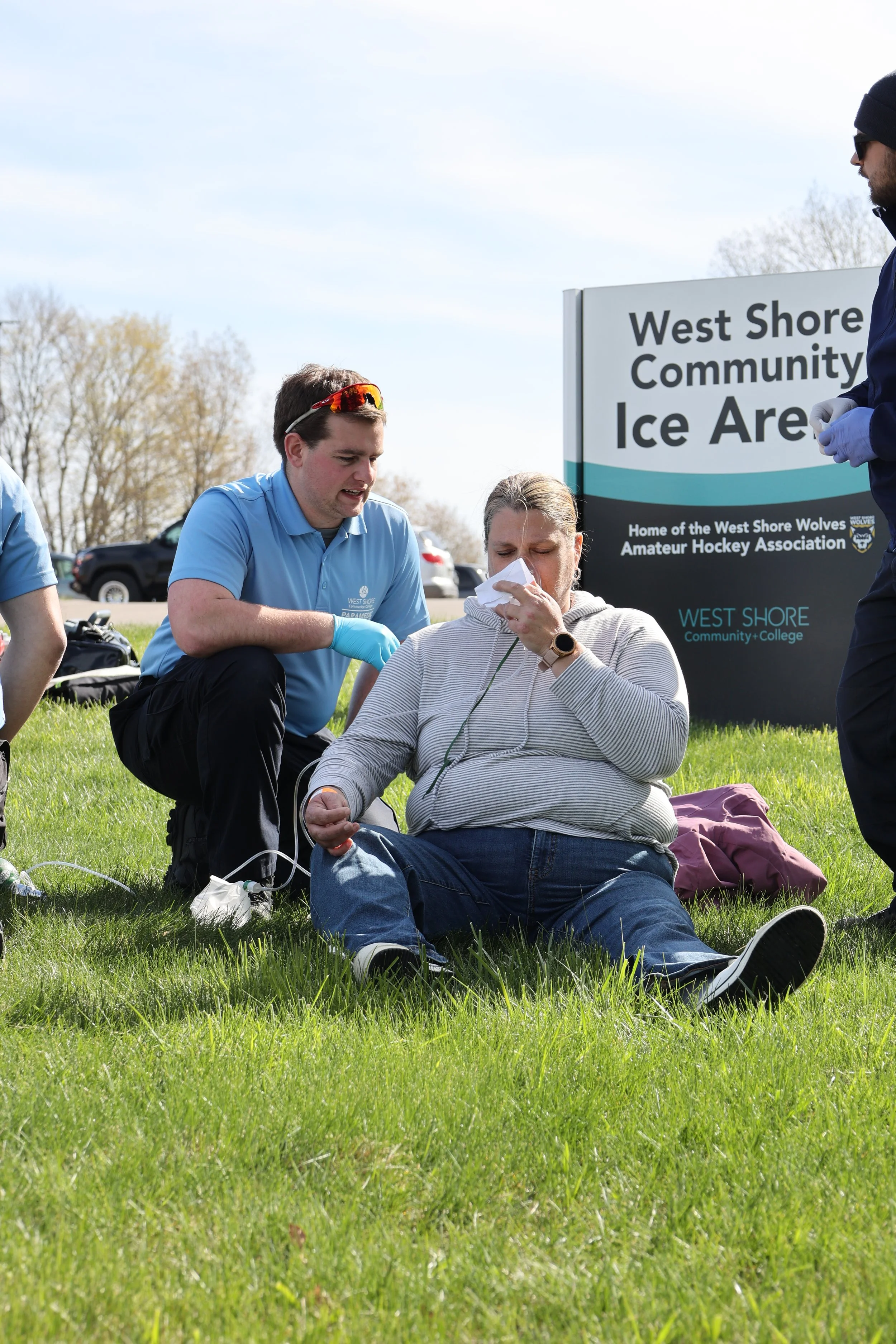 Emergency responders assist a person outside West Shore Community Ice Arena. The person is sitting on grass, holding a mask to their face, while a responder in a blue uniform kneels beside them, providing aid.