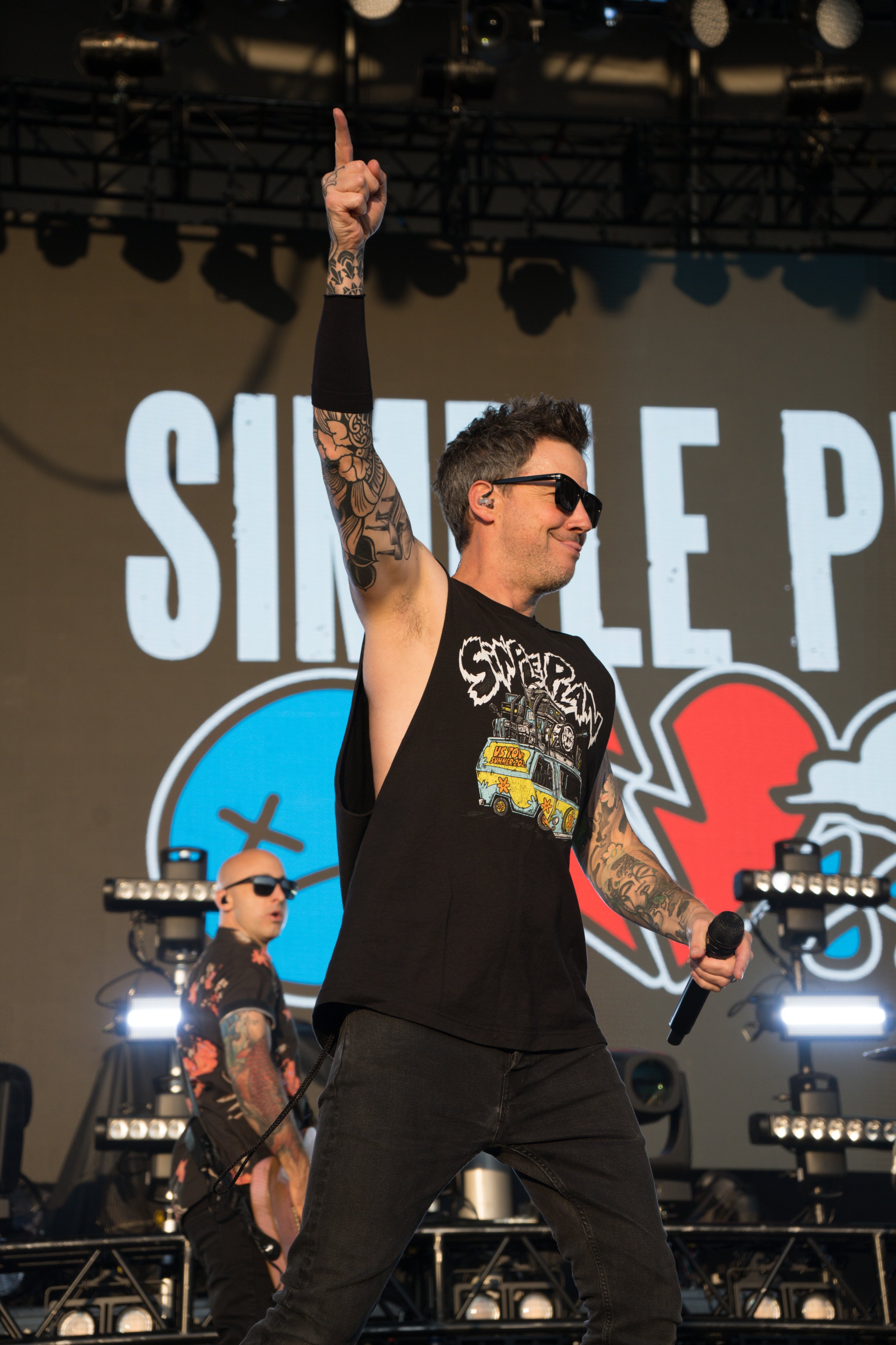 Musician performing on stage in front of "Simple Plan" backdrop, wearing sunglasses and a tank top with tattoos on arm.