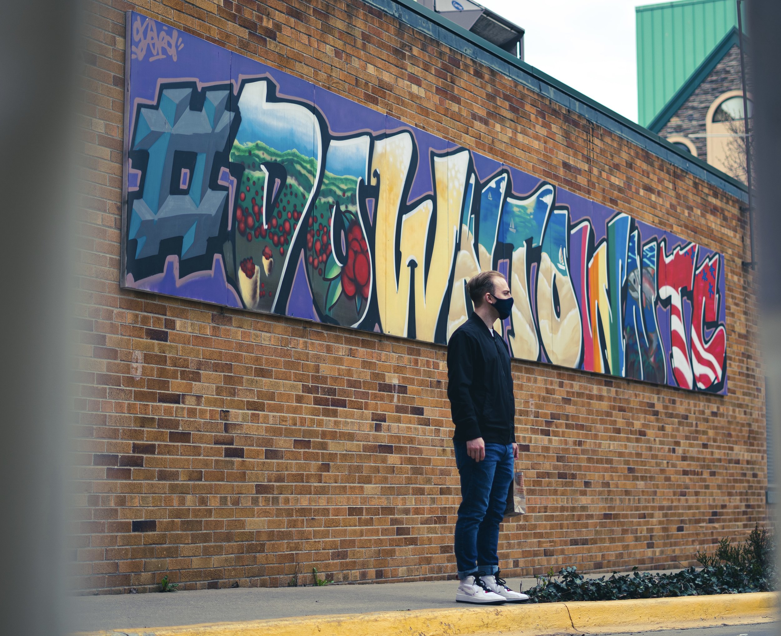 A man wearing a mask stands in front of a colorful graffiti mural on a brick wall.
