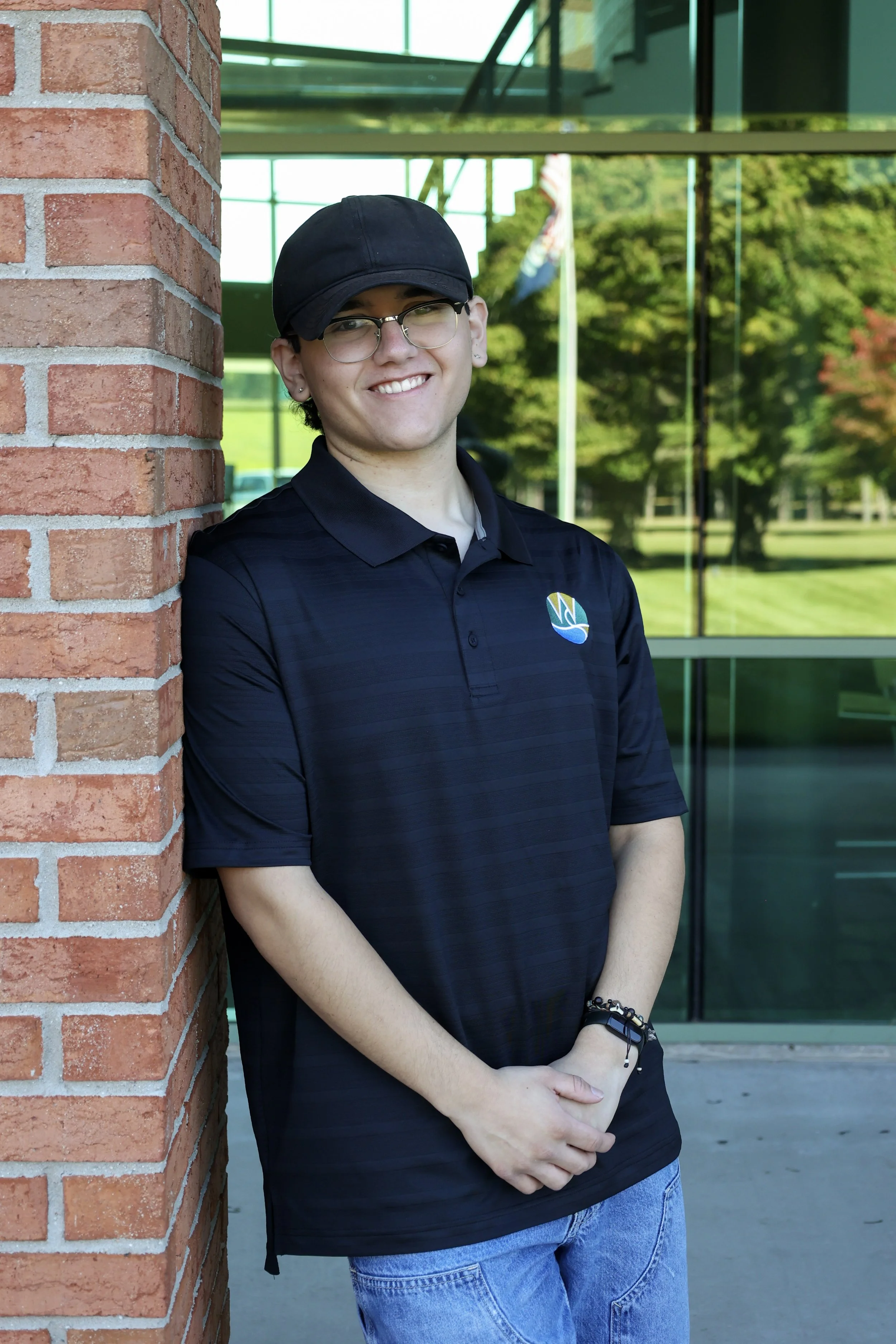 Person leaning against a brick wall, wearing a black cap, glasses, and a black polo shirt with a logo, standing in front of a glass window.