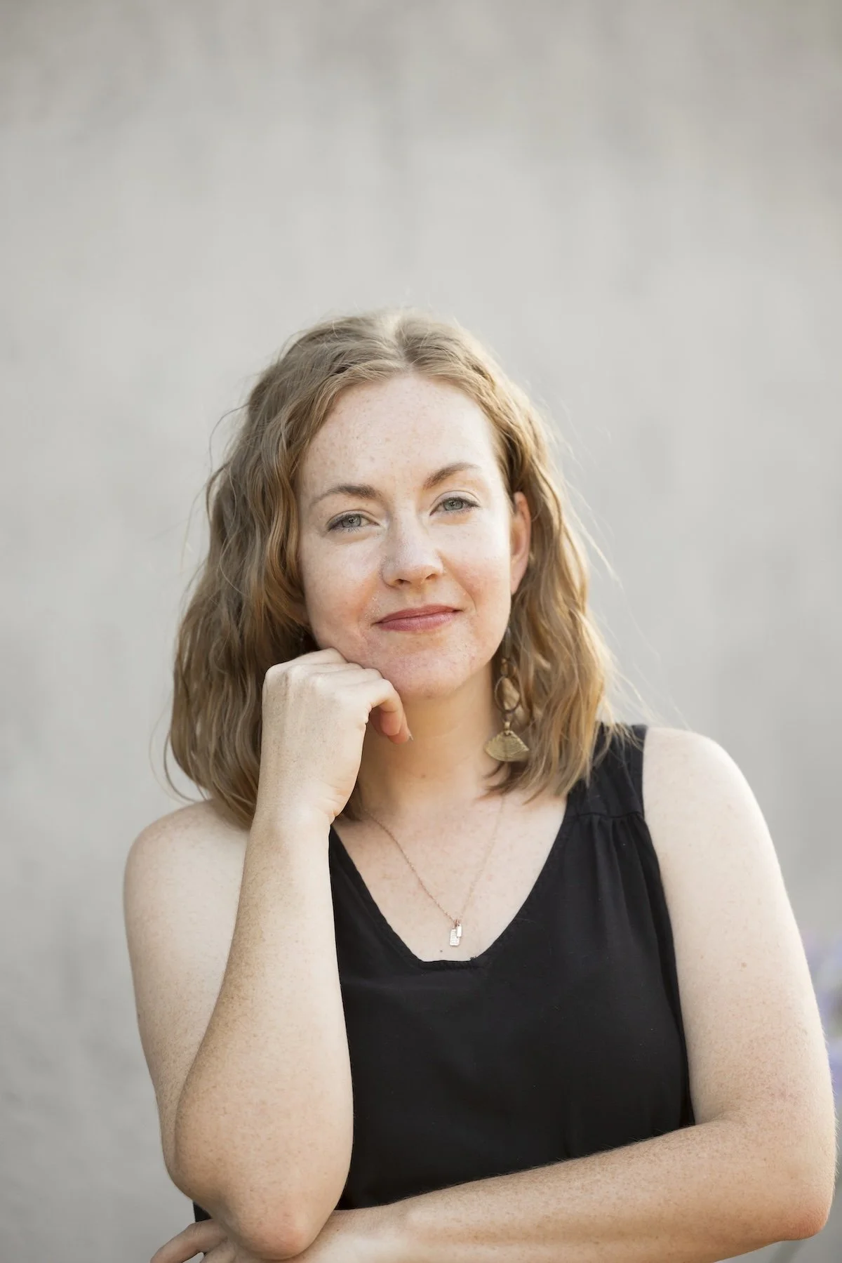 A woman with shoulder-length wavy red hair and fair skin, wearing a black sleeveless top, gold earrings, and a delicate necklace, smiling softly with her chin resting on her hand kept under her chin, standing against a blurred gray background.