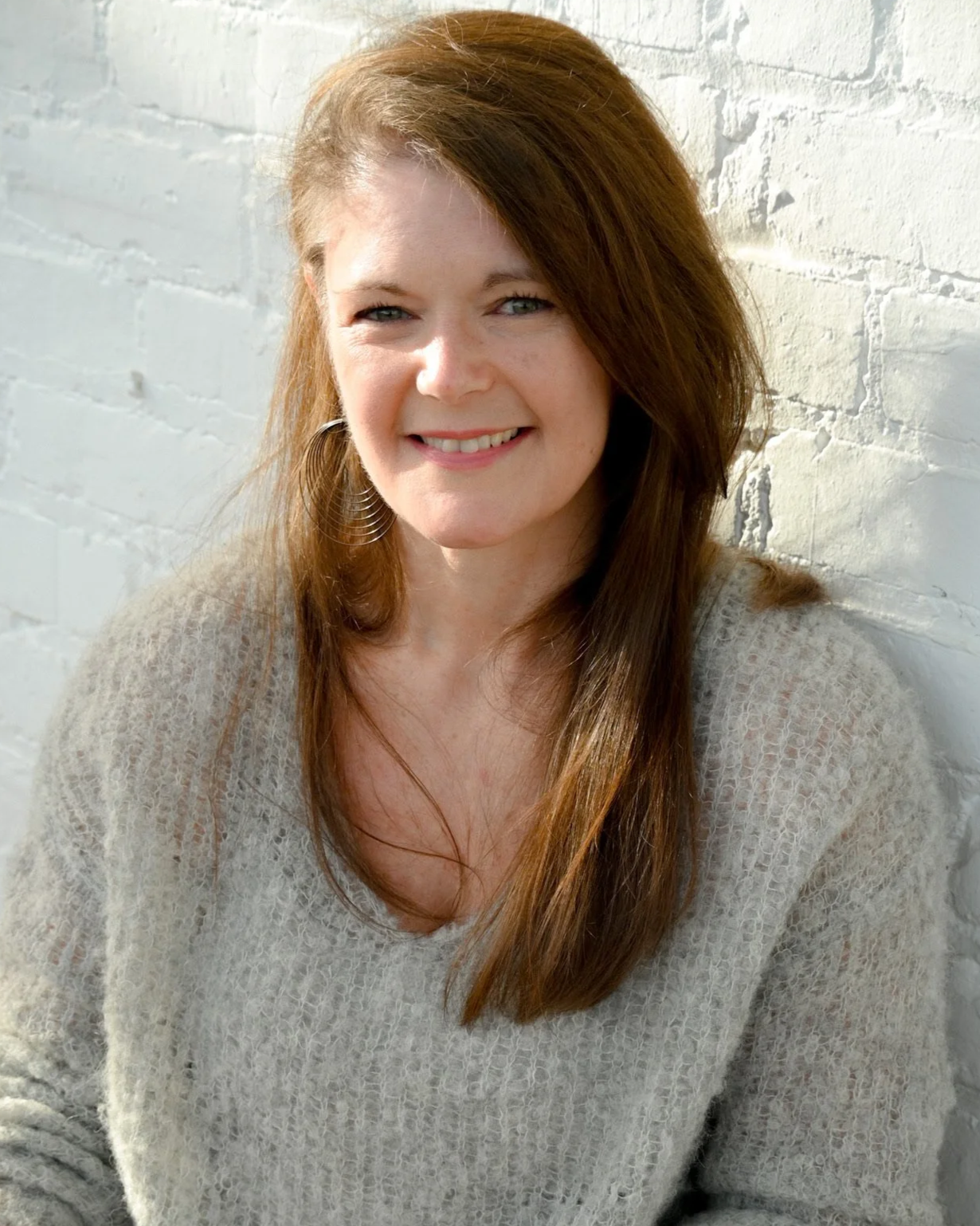 A woman with long reddish-brown hair smiling, wearing a light beige sweater, standing against a white brick wall in sunlight.
