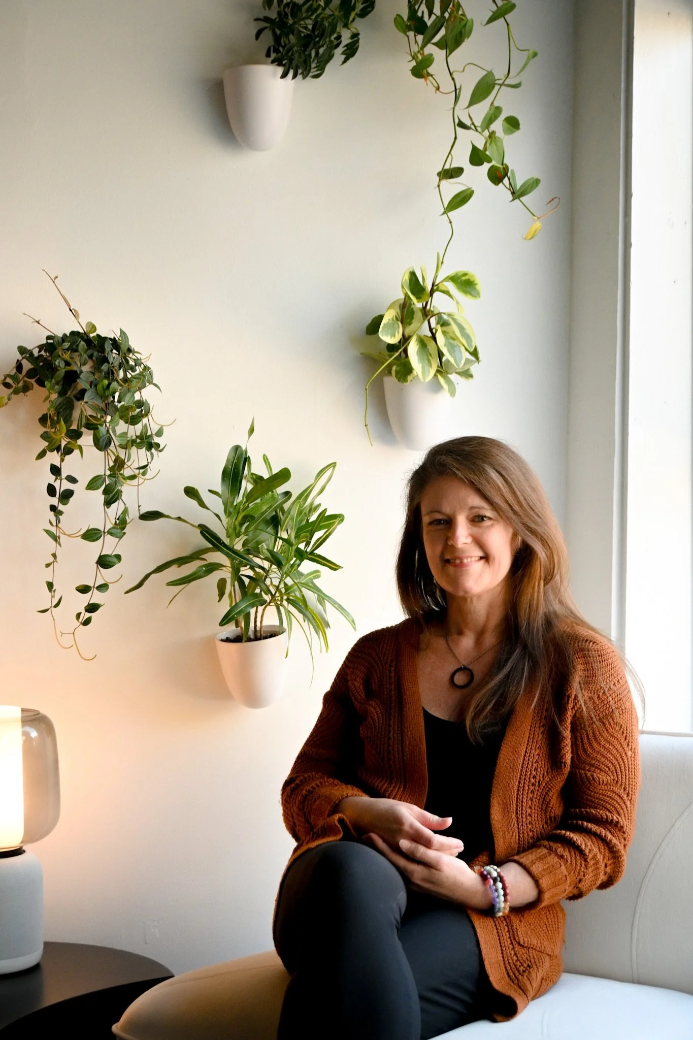 A woman sitting on a white chair by a window with potted plants on the wall behind her.